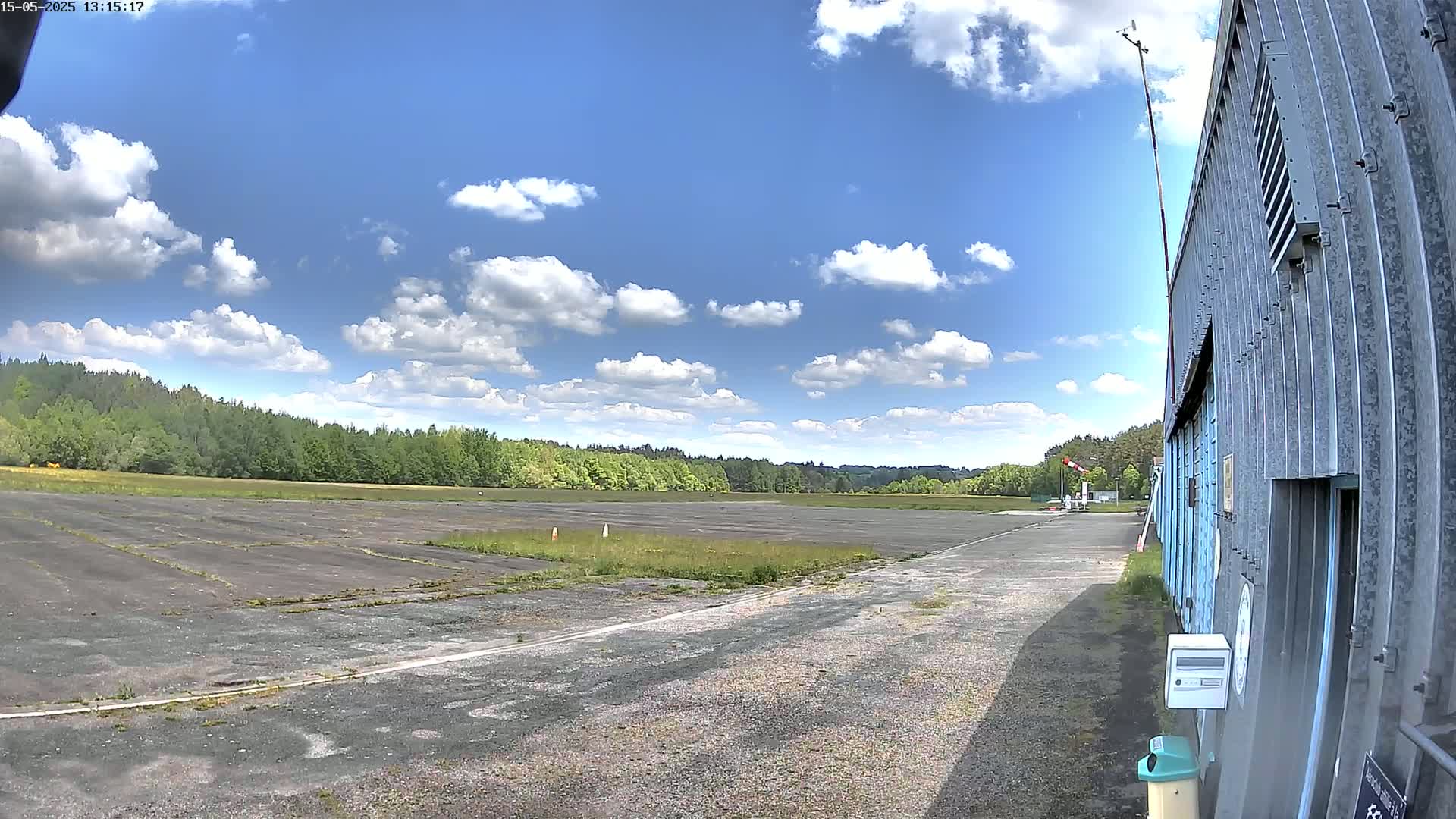 A mostly sunny day with scattered cumulus clouds shows a view of a paved airfield next to a corrugated metal building, with a line of trees in the background.