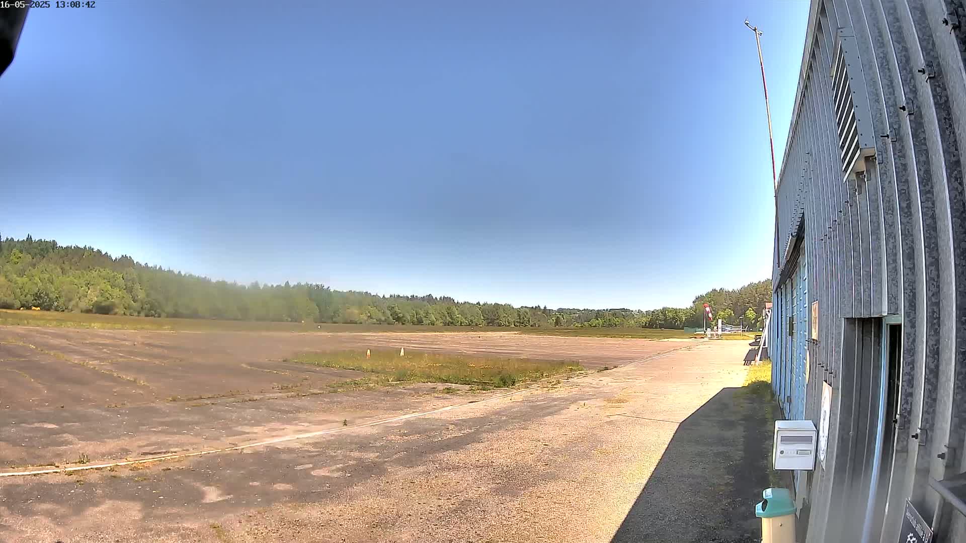 A mostly clear blue sky overlooks a flat, empty airfield next to a corrugated metal building on a sunny day.