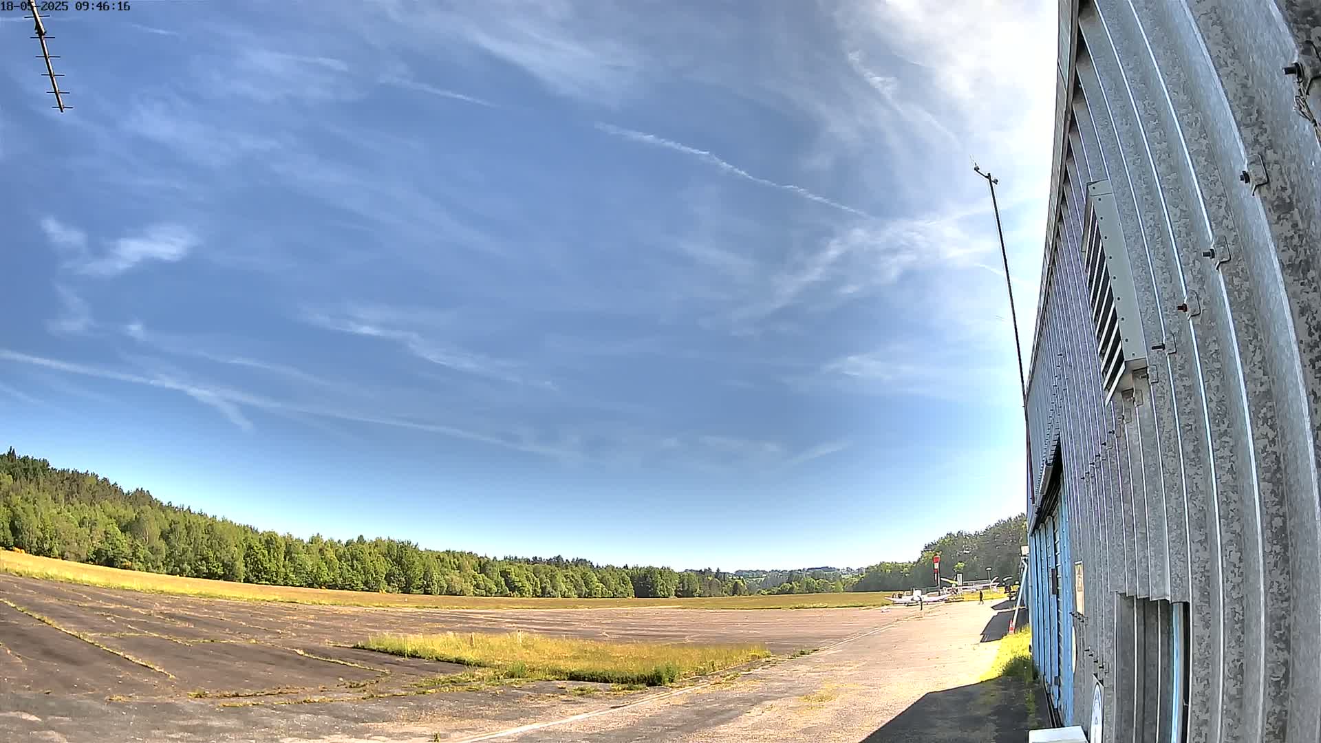 A mostly sunny day with scattered clouds above a small airfield next to a corrugated metal building.