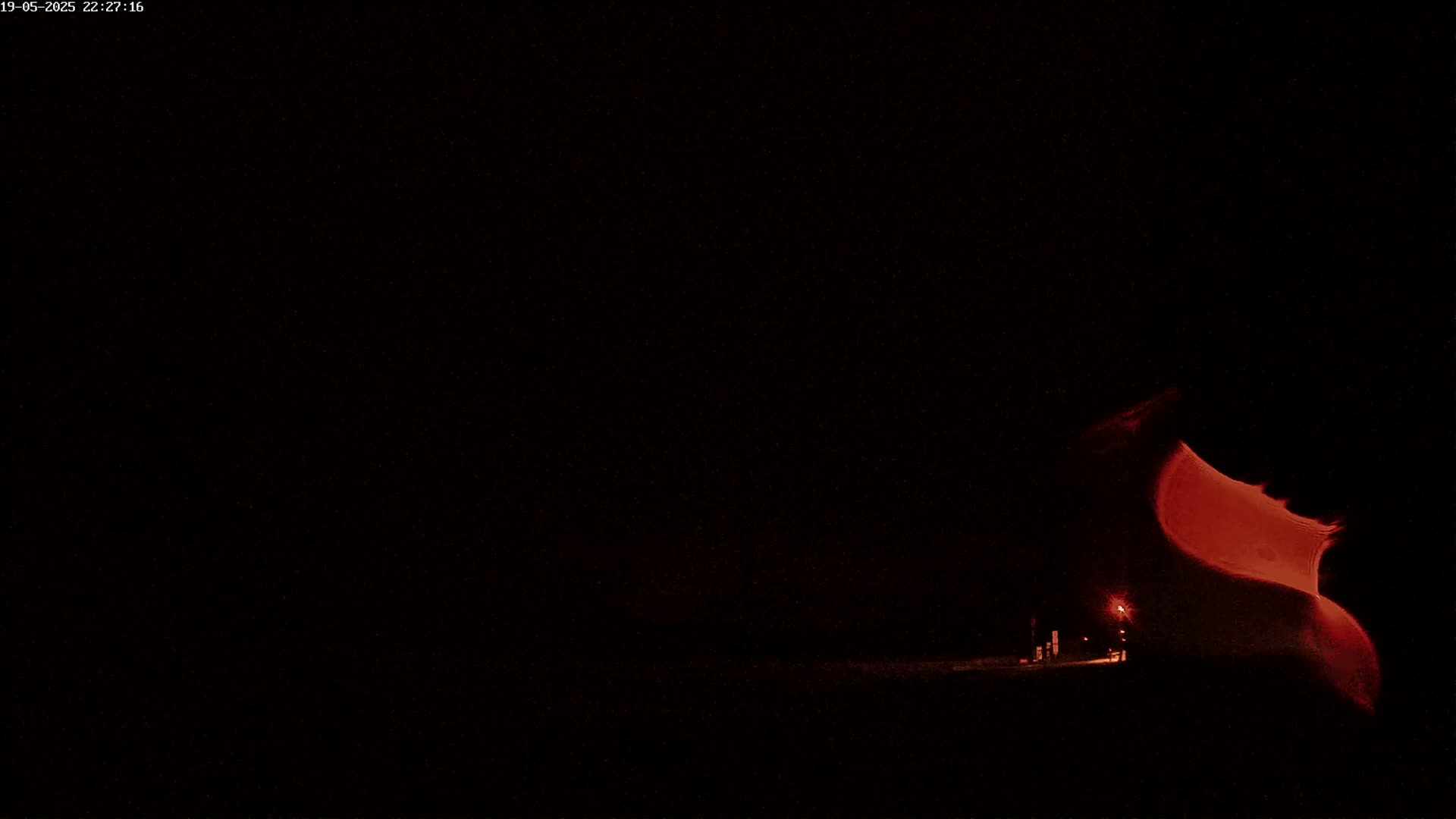 A dimly lit, dark landscape at night shows a red glow emanating from a figure in the right corner, with faint lights visible in the distance.
