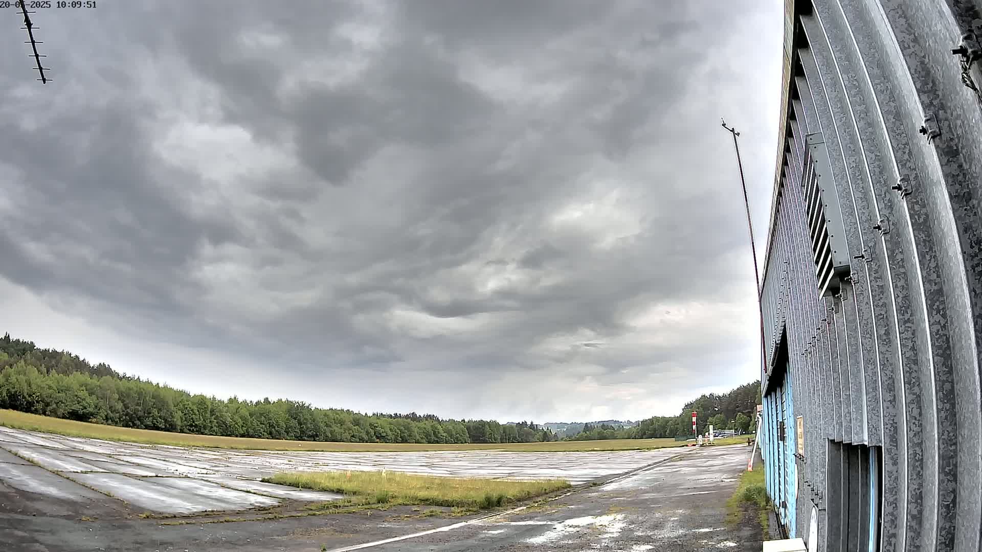 A mostly cloudy sky hangs over a paved, empty airfield bordered by trees and a corrugated metal building.