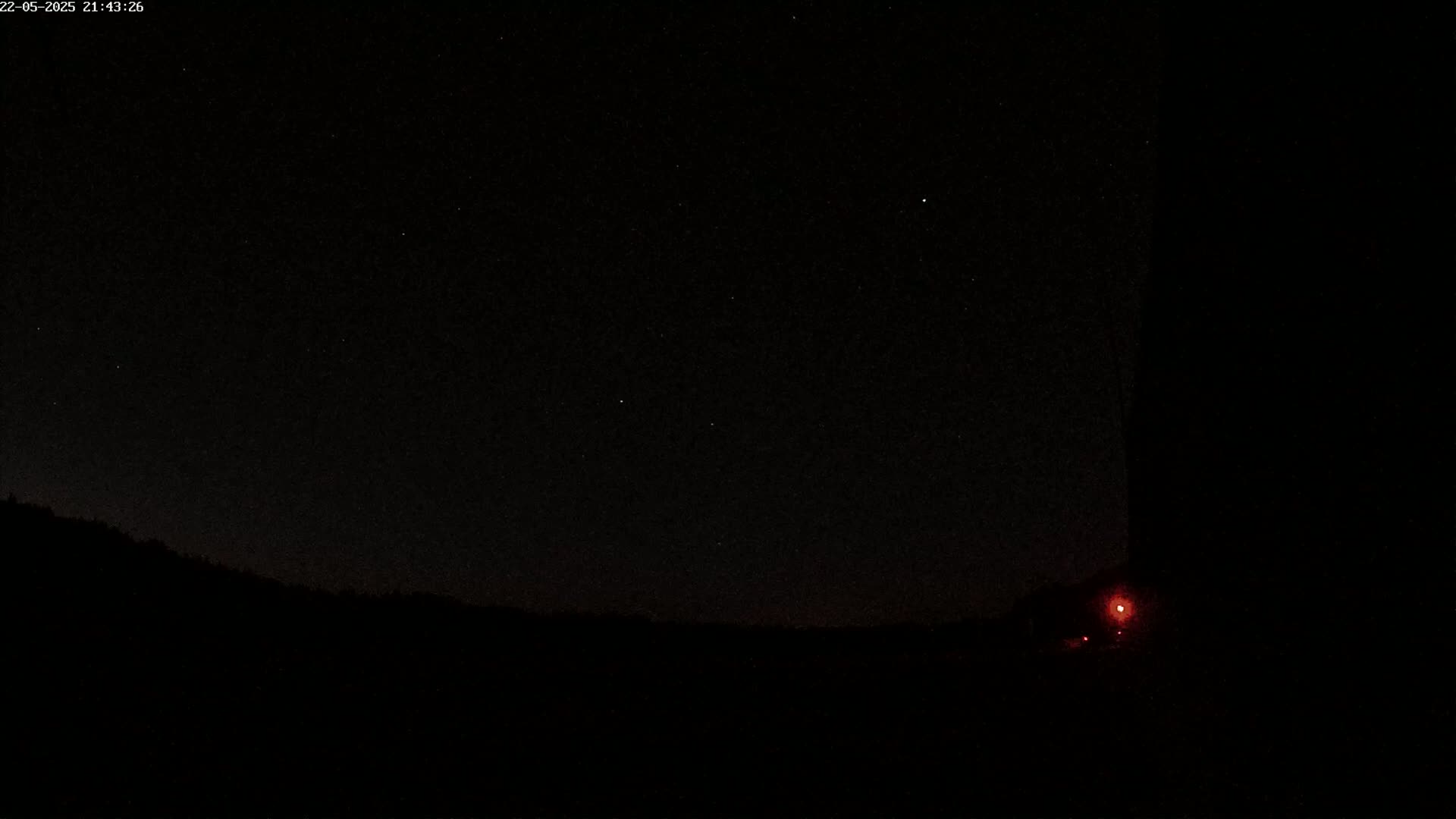 A nighttime landscape shows a dark silhouette of hills under a clear, starry sky with a small red light visible near the bottom right corner.