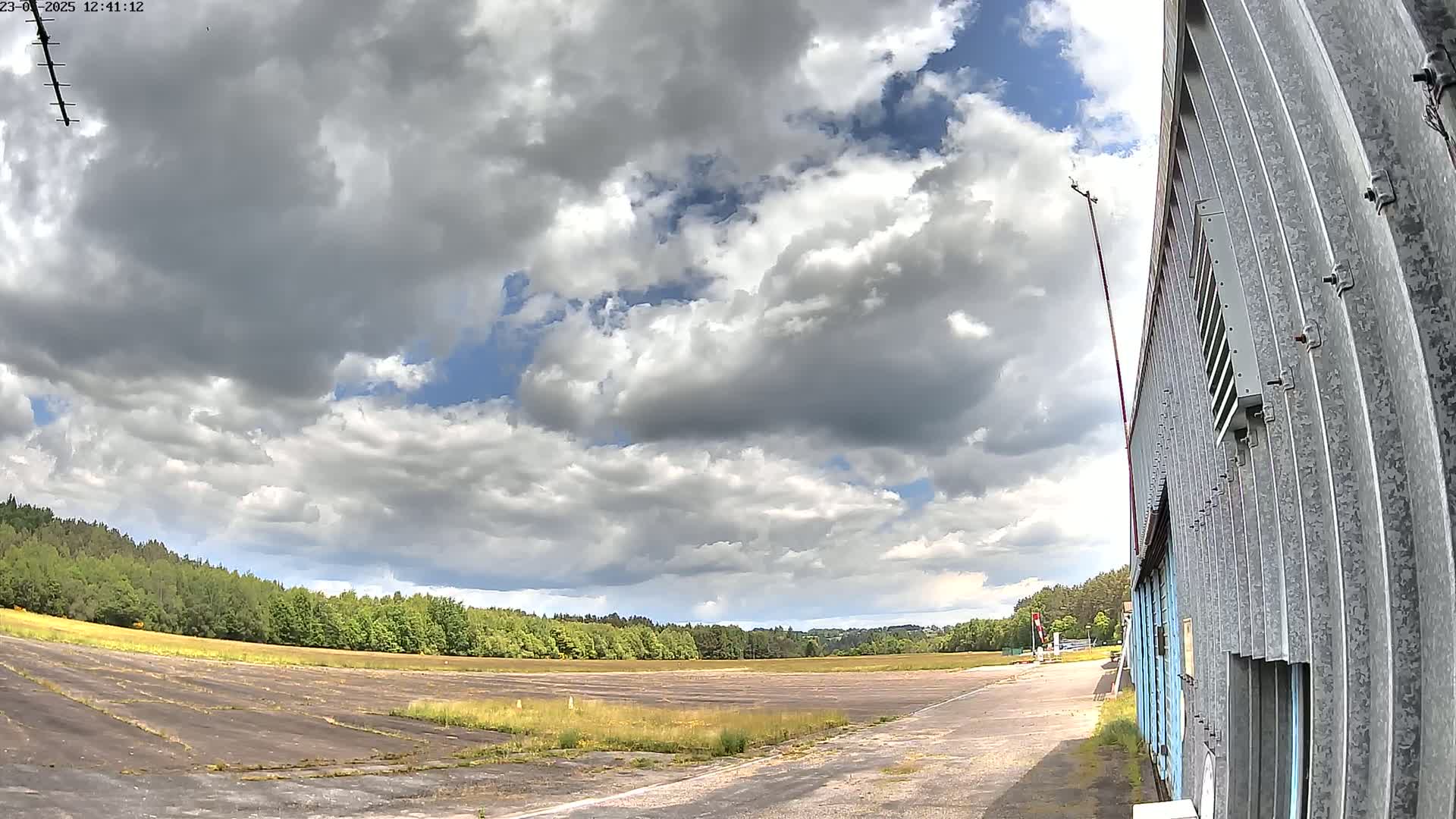 A partly cloudy sky overlooks a grassy airfield next to a corrugated metal building.