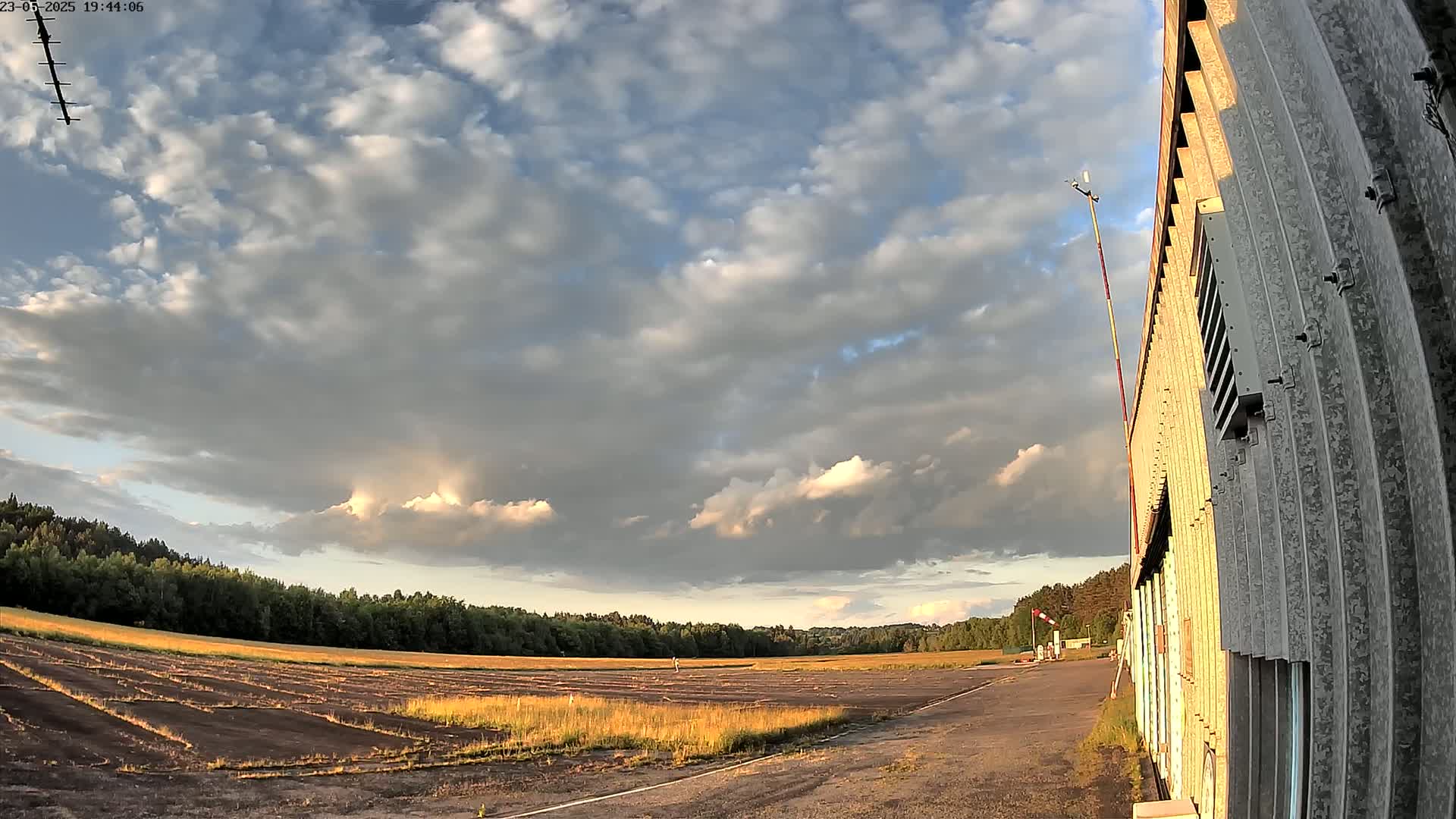 A partly cloudy sky over a flat, grassy airfield next to a corrugated metal building.