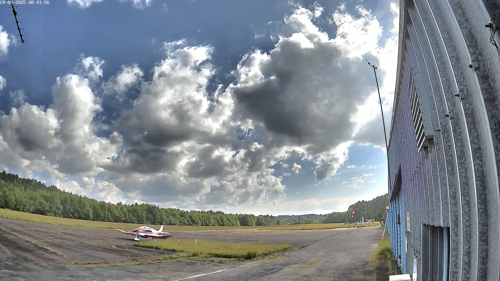 A small, red and white airplane sits on a tarmac next to a building under a partly cloudy sky.
