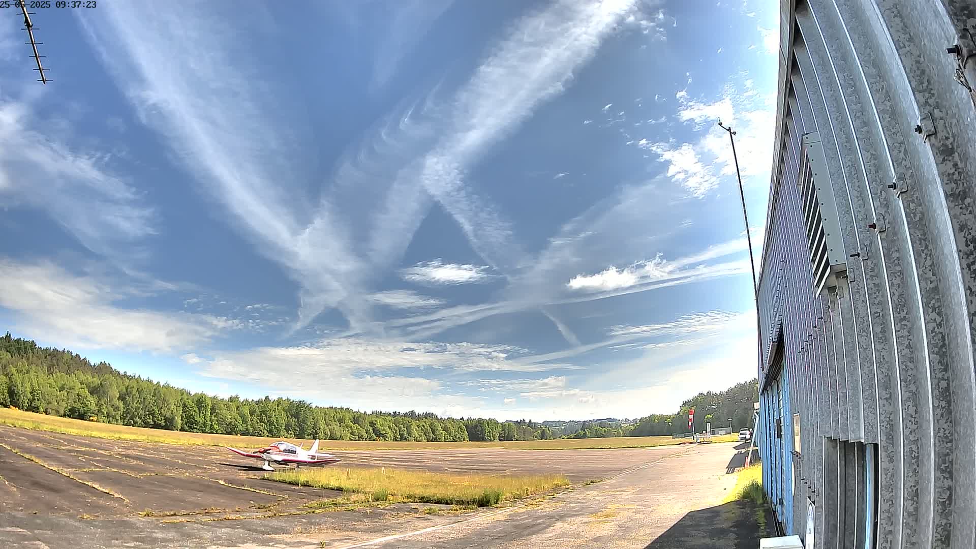 A small airplane sits on a tarmac next to a building under a mostly sunny sky with scattered clouds and contrails.
