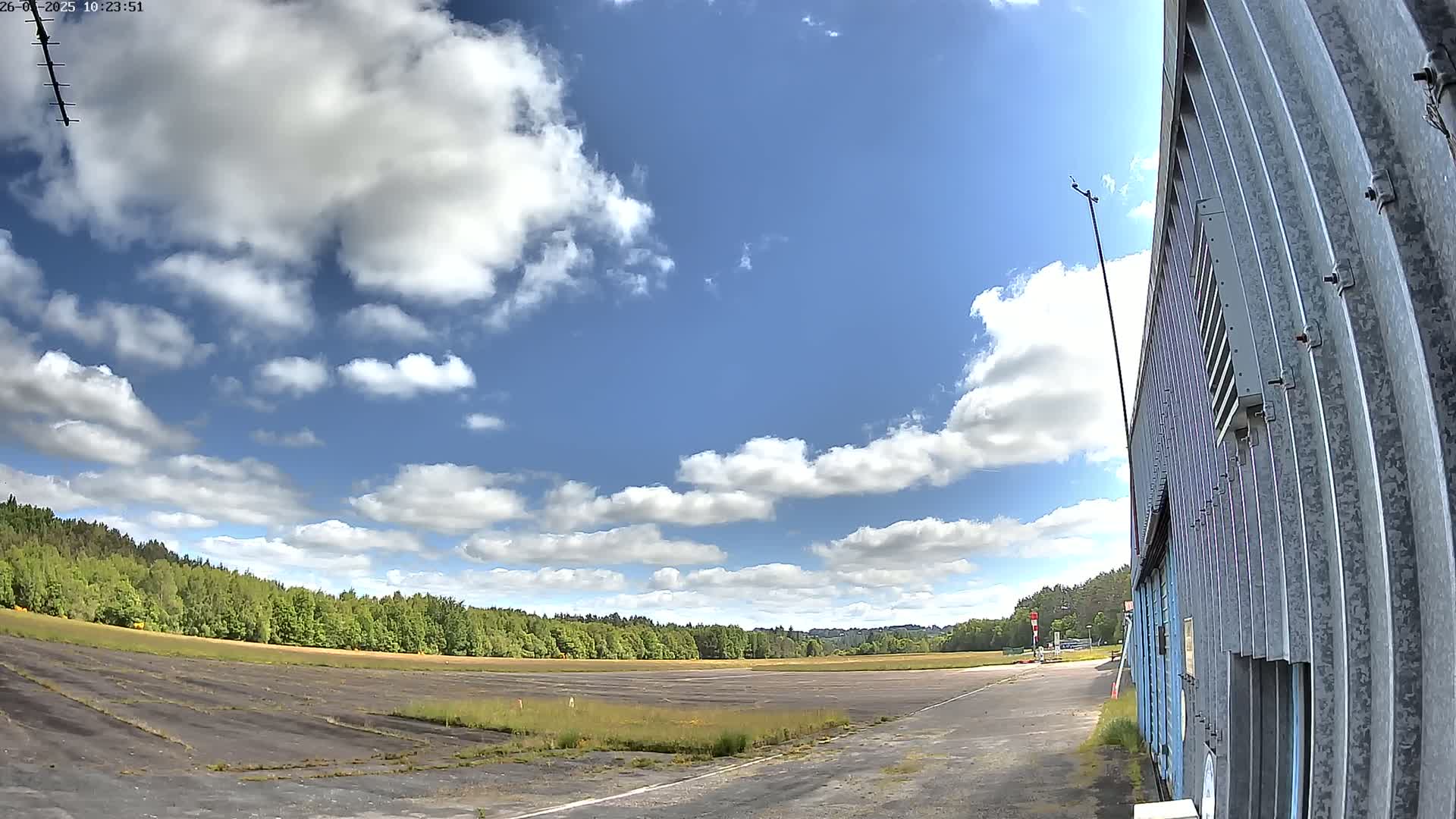 A mostly sunny day with scattered clouds over a paved field bordered by trees and a corrugated metal building.