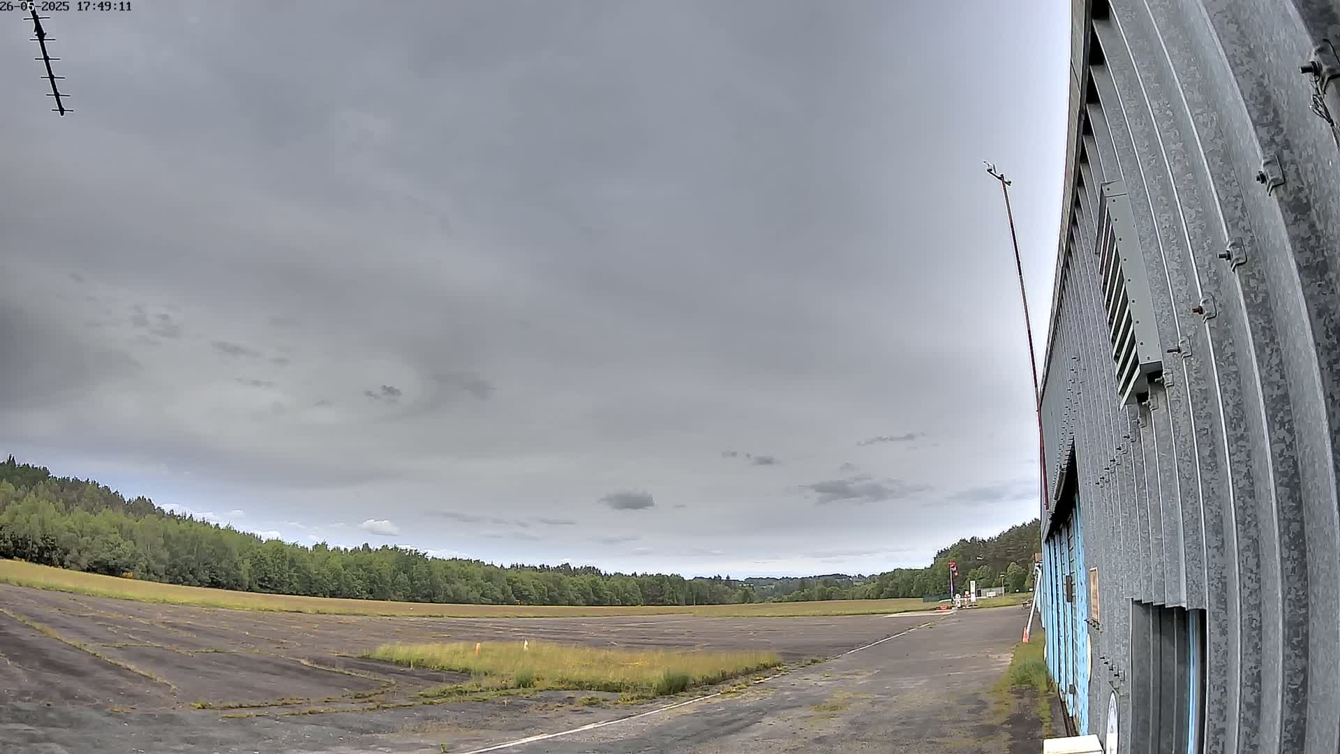 A mostly overcast sky hangs over a flat, paved area next to a long, corrugated metal building, with a line of trees in the distance.