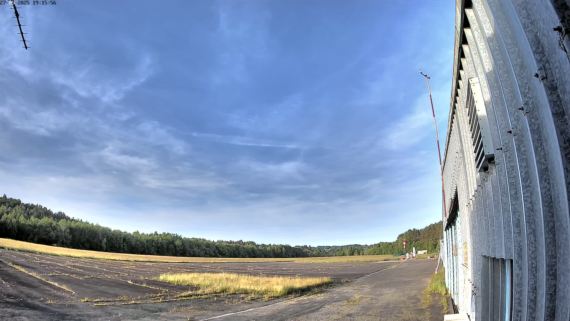 A mostly sunny sky with thin clouds hangs over a flat, grassy field bordered by trees and a weathered metal building.