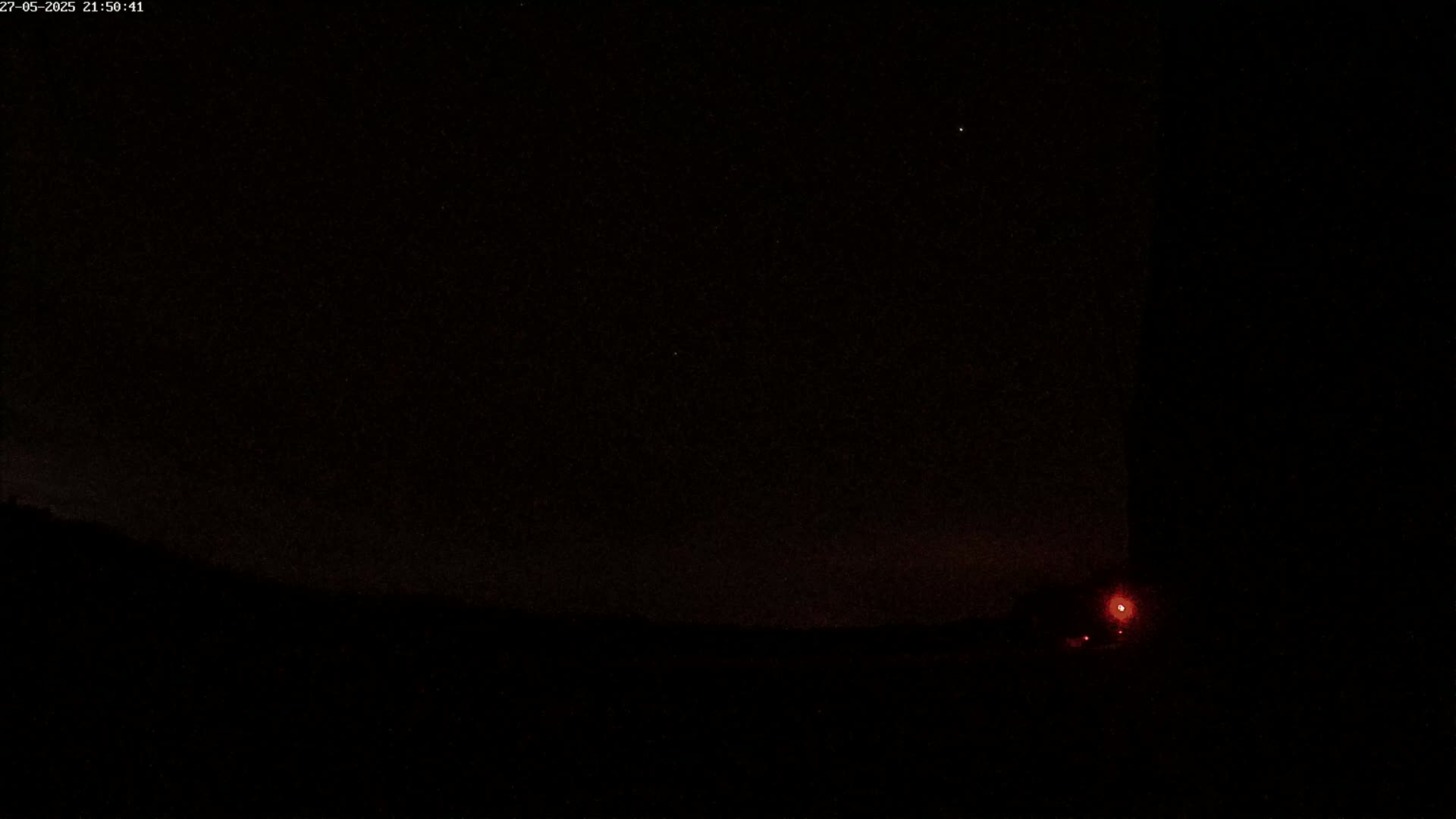 A dark, nighttime scene shows a star-filled sky above a silhouetted landscape with a single red light visible near the bottom right.