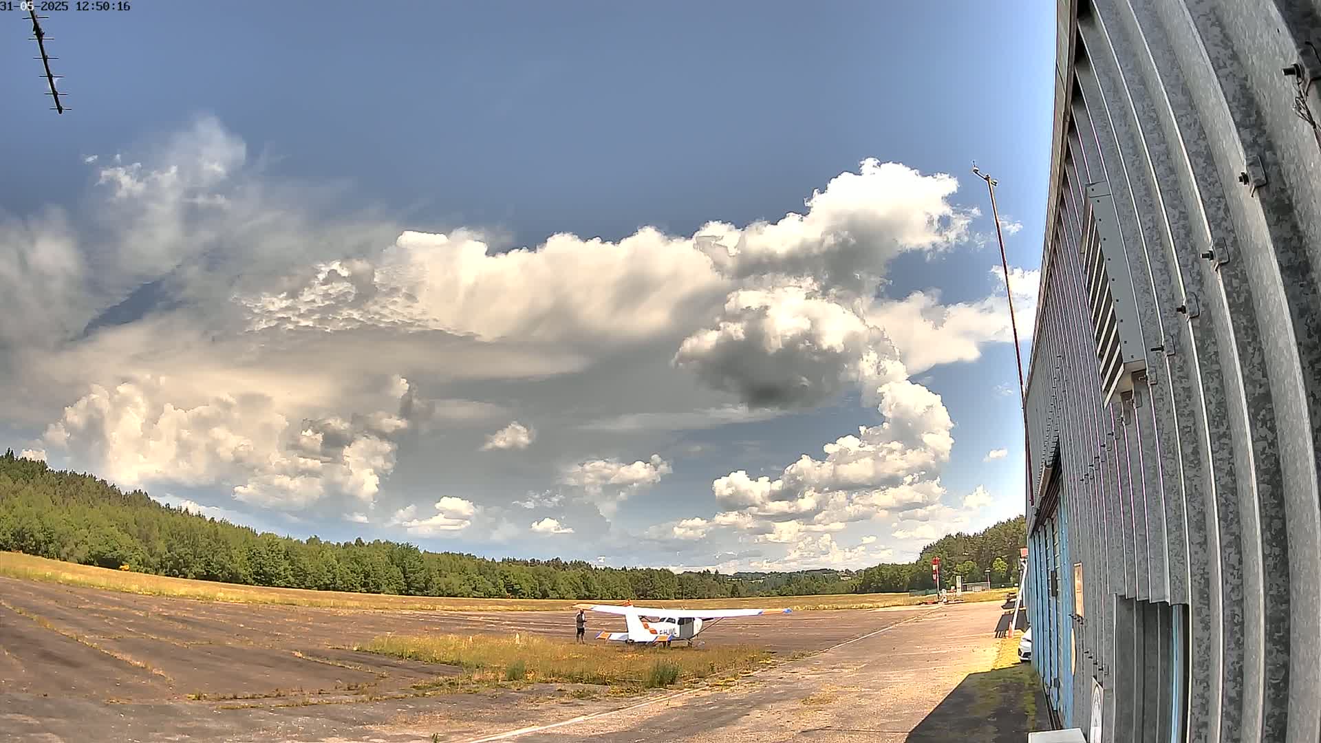 A small airplane sits on a tarmac next to a hangar under a partly cloudy sky.