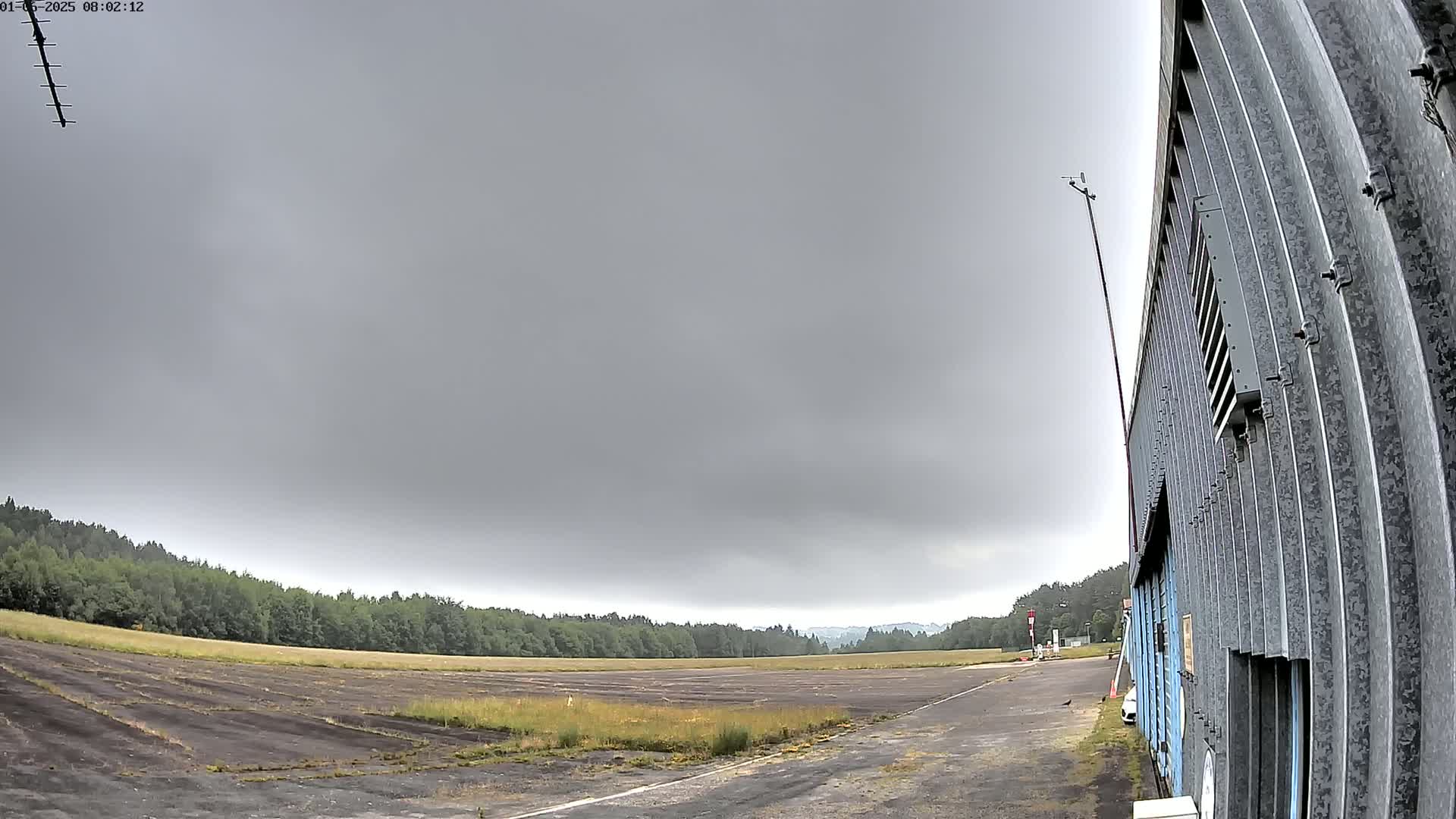 A large, corrugated metal building stands next to a long, empty airfield under a cloudy, overcast sky.