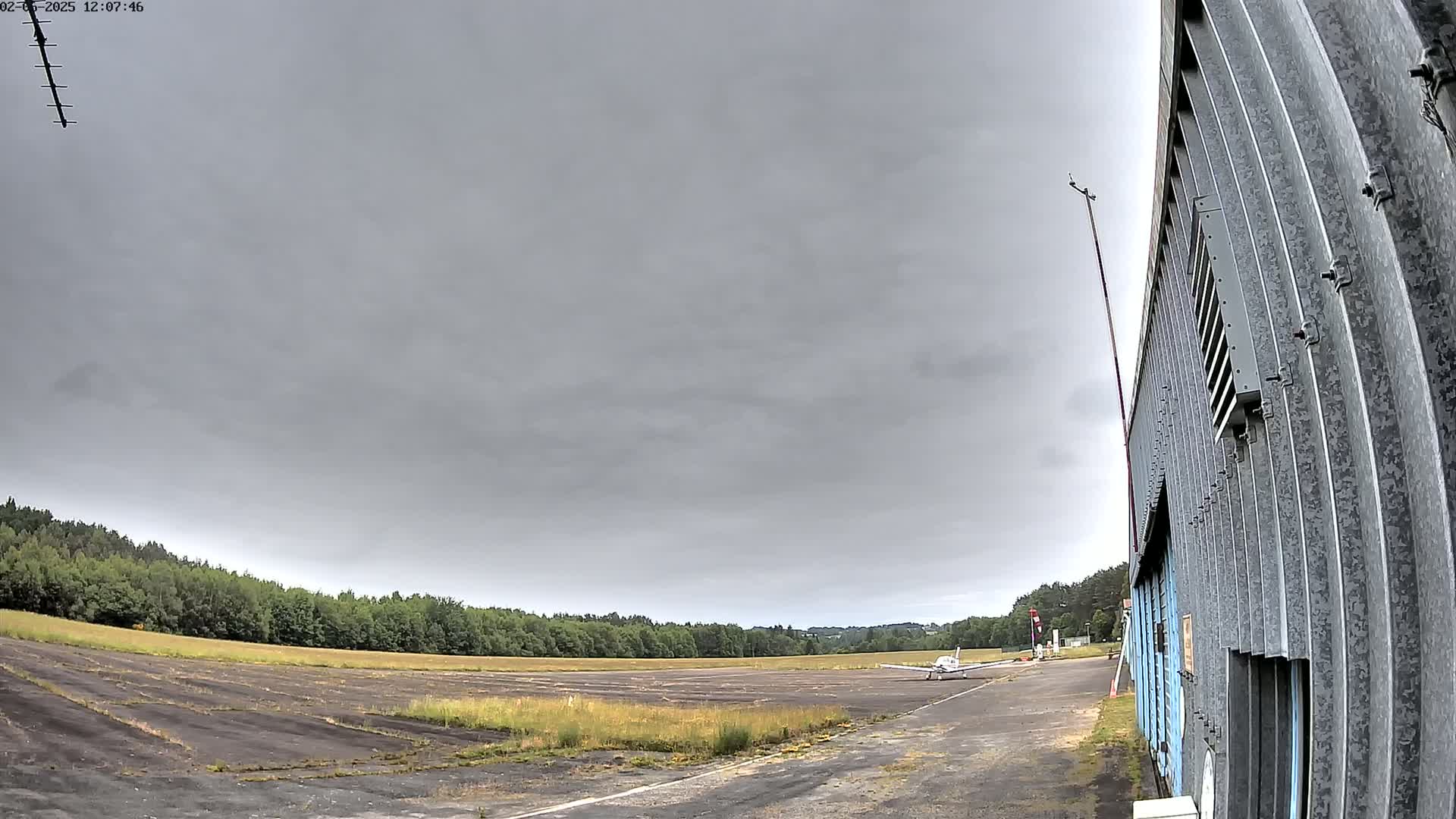 A small airplane sits on a tarmac next to a corrugated metal hangar under an overcast sky.