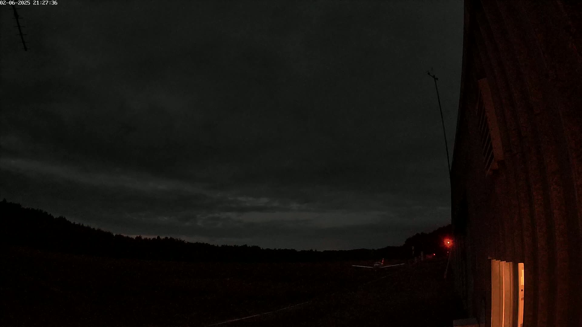 A dark, overcast night scene shows a silhouetted treeline and a building with a lit interior, under a mostly cloudy sky.