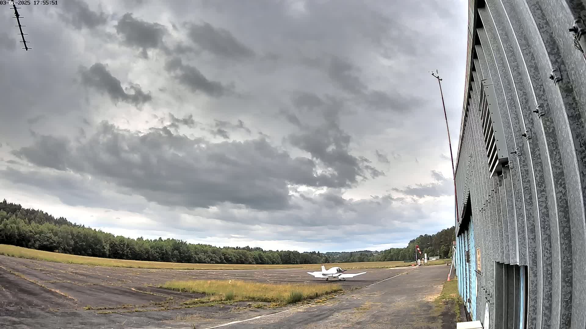 A small white airplane sits on a tarmac next to a corrugated metal building under a mostly cloudy sky.