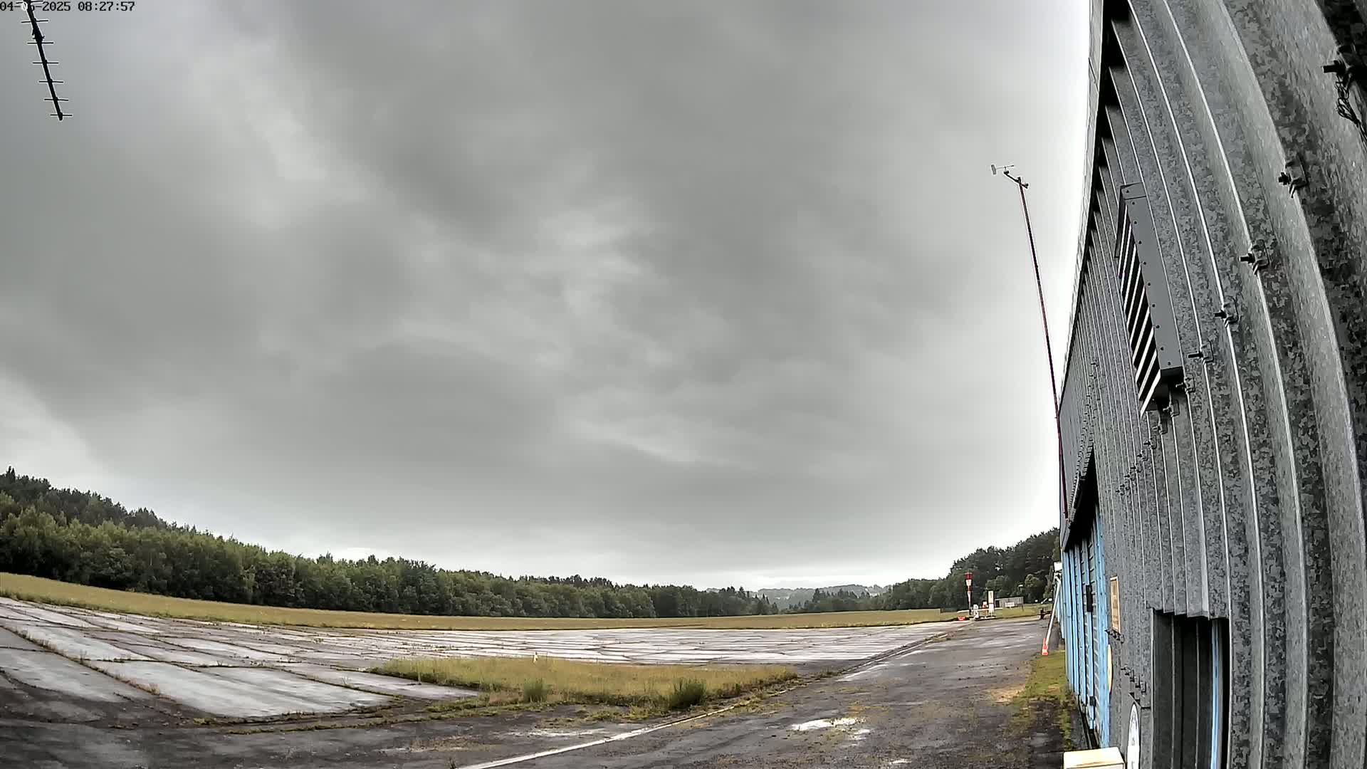 A disused airfield under a cloudy sky is partially visible, next to a corrugated metal building.
