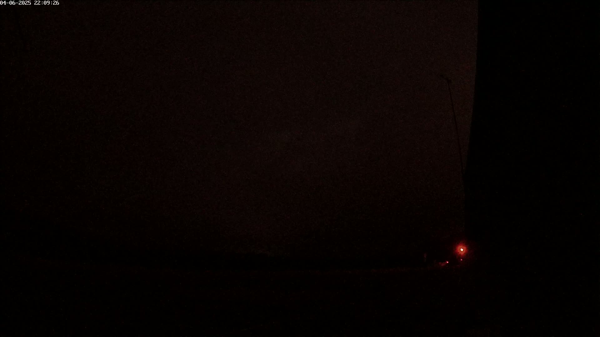 A dark, nighttime scene shows a dimly lit roadway with a single red light visible in the distance.