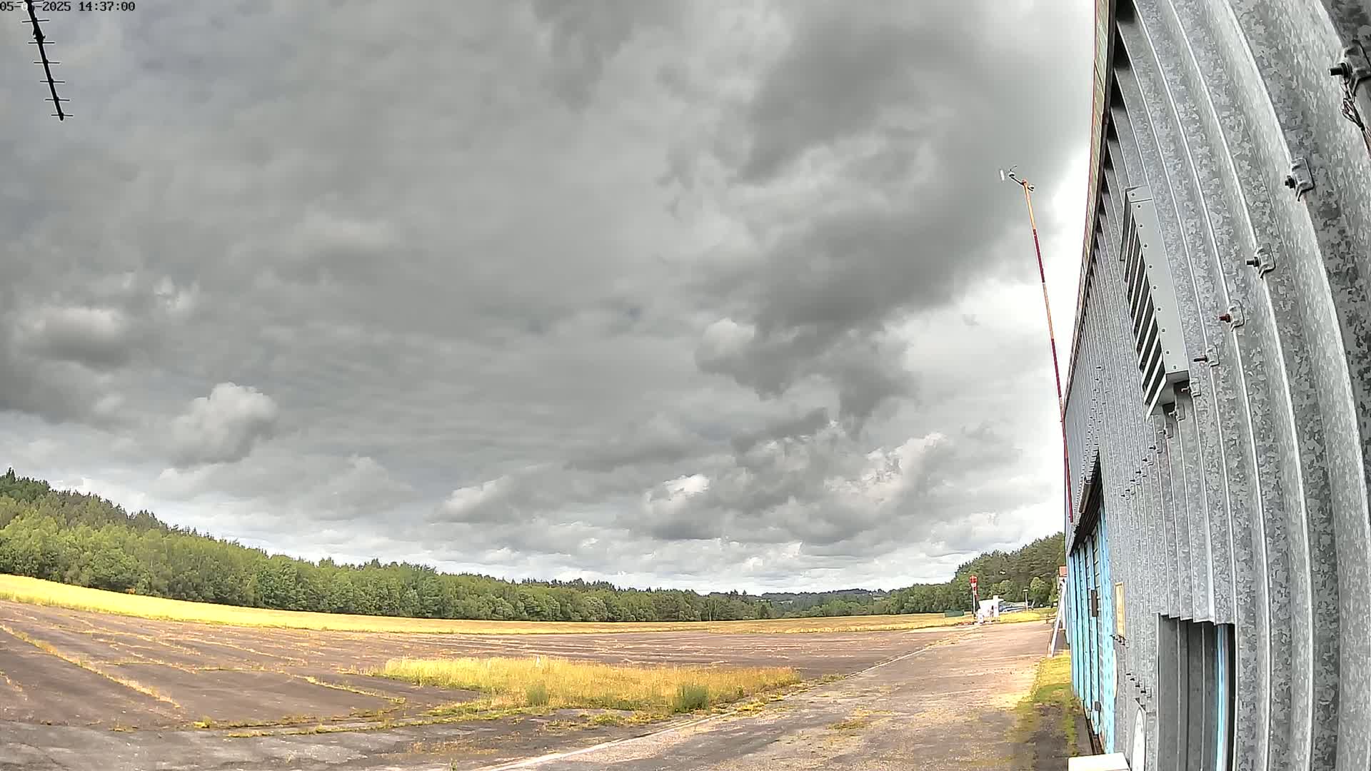 A wide, overcast sky looms over a flat, grassy field next to a corrugated metal building.