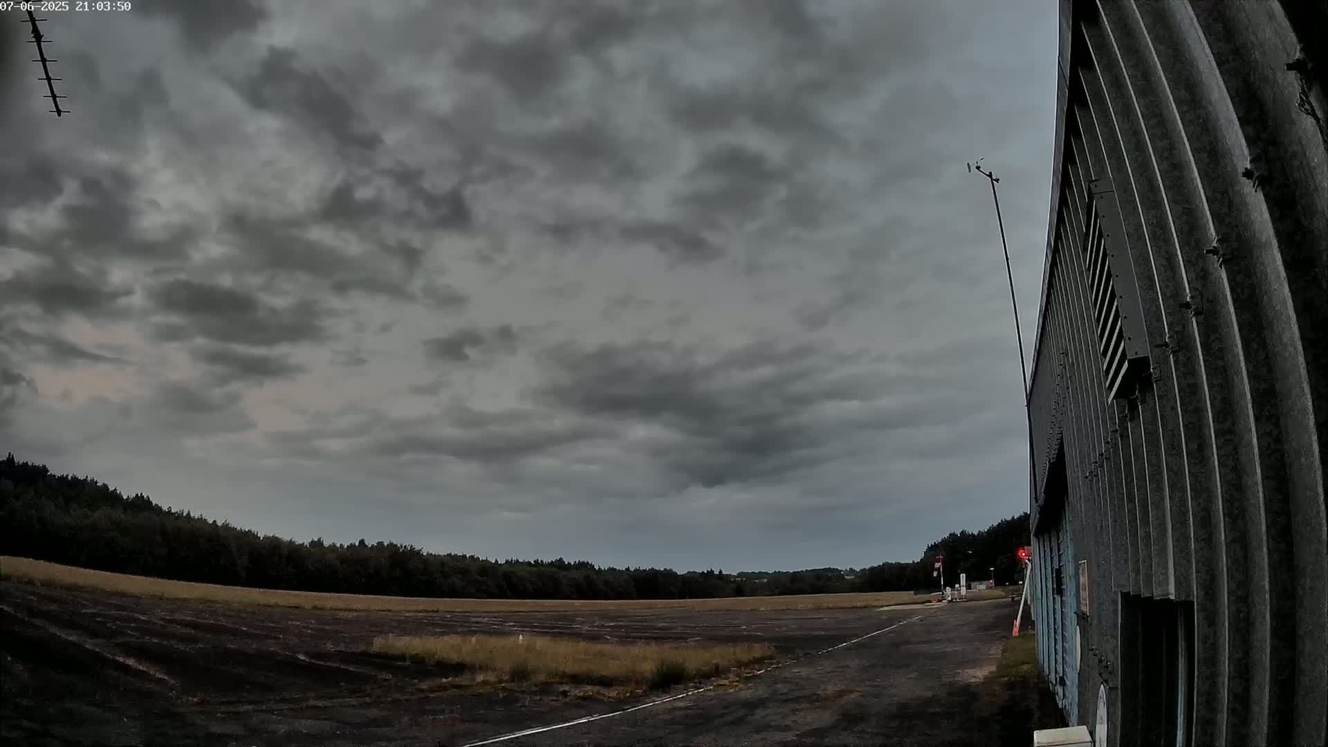 A dark, overcast sky looms over a flat, paved area next to a corrugated metal building and a line of trees.