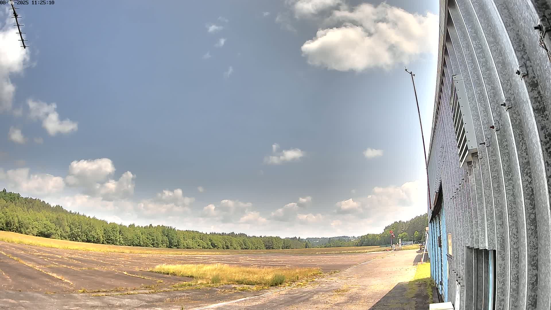 A partly cloudy sky overlooks a flat, overgrown field next to a corrugated metal building.