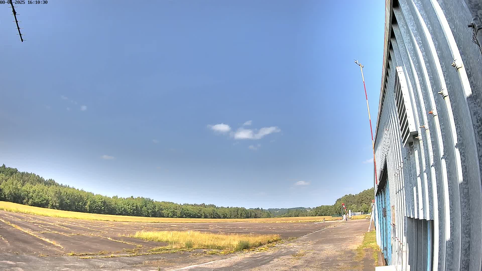 A mostly clear blue sky overlooks a disused airfield with overgrown grass and a corrugated metal building.