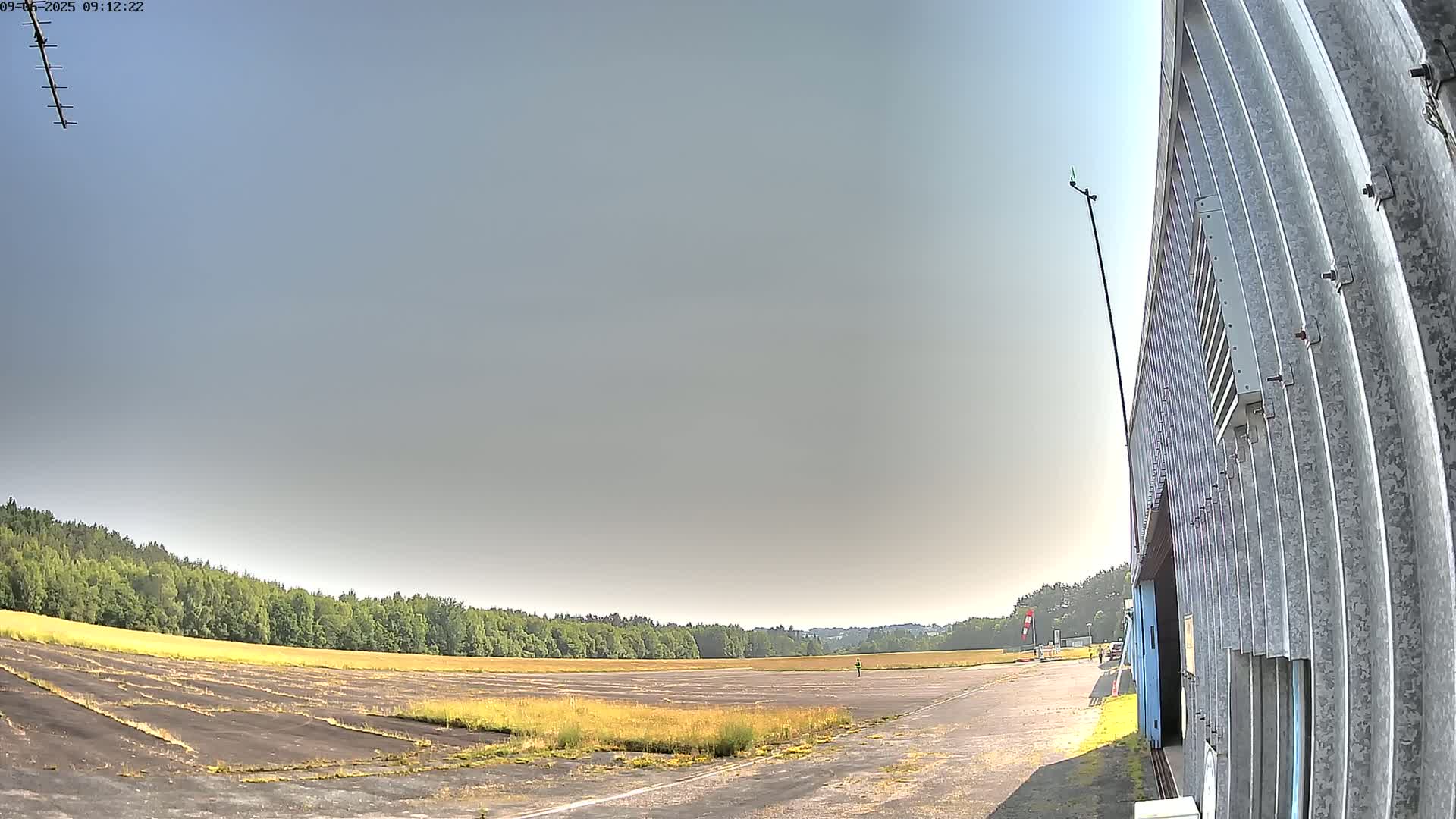 A mostly sunny day reveals an airfield with overgrown grass and a large corrugated metal building.
