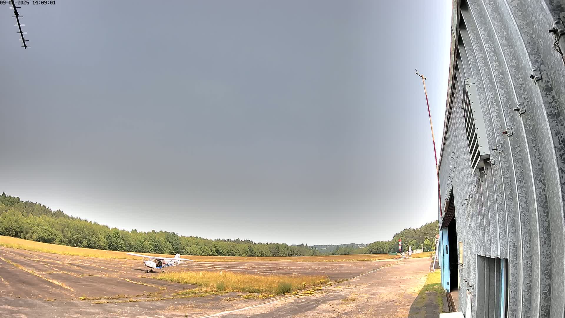 A small airplane sits on a grassy airfield next to a corrugated metal hangar under a mostly clear sky.