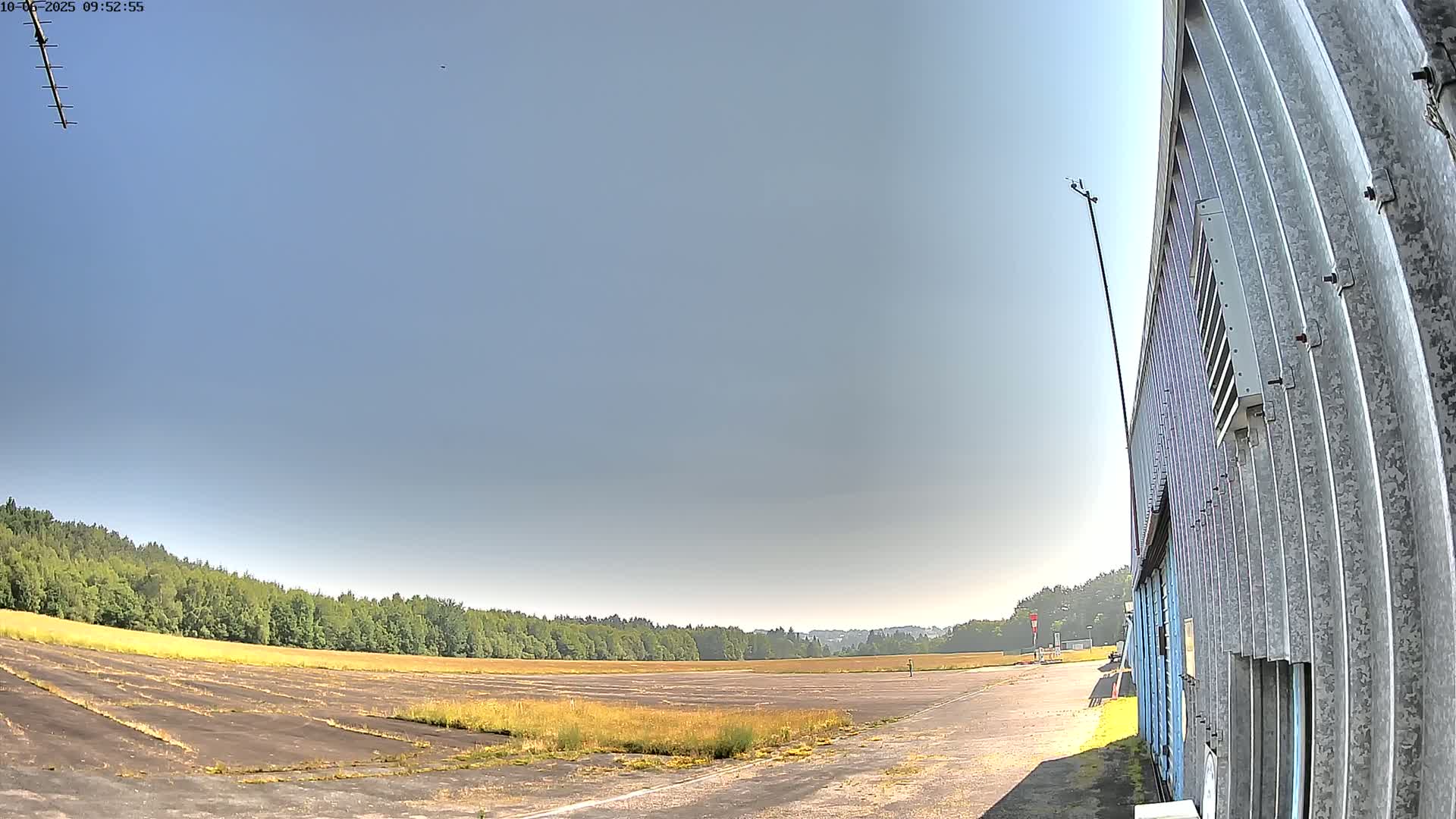 A large, empty tarmac area bordered by trees under a clear, sunny sky is partially visible next to a corrugated metal building.