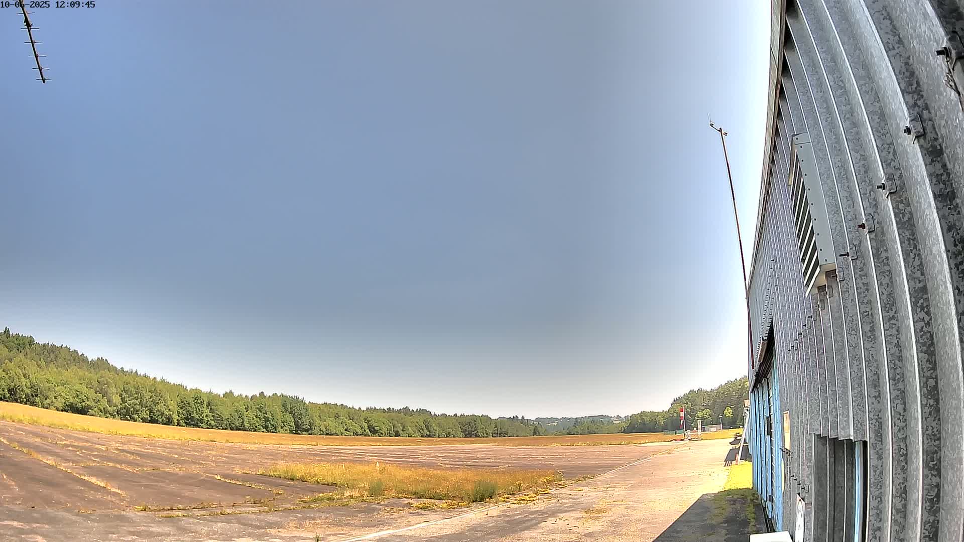 Under a clear blue sky, a wide, empty, paved area, possibly an airfield, stretches toward a line of trees, next to a corrugated metal building.
