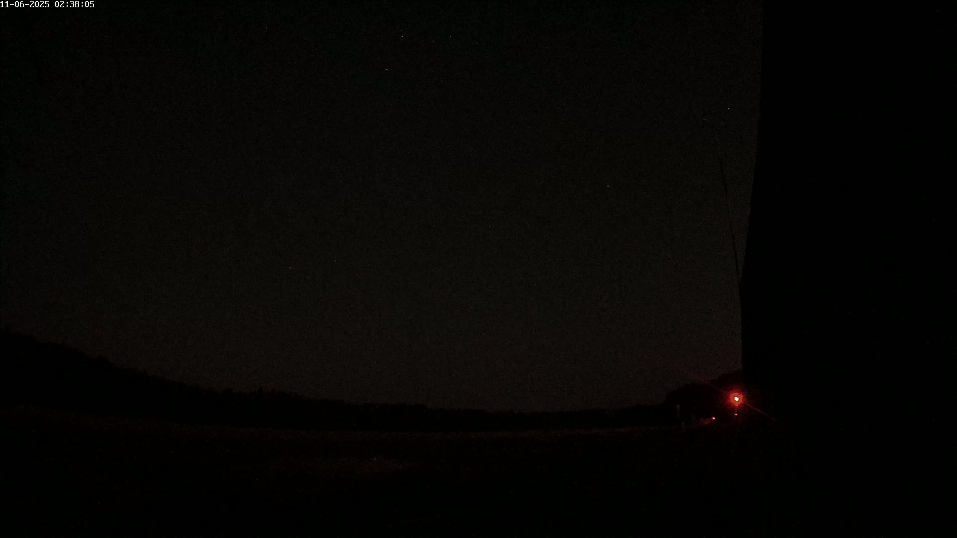 A dark, nighttime scene shows a silhouetted landscape under a starry sky with a faint red light visible in the lower right corner.