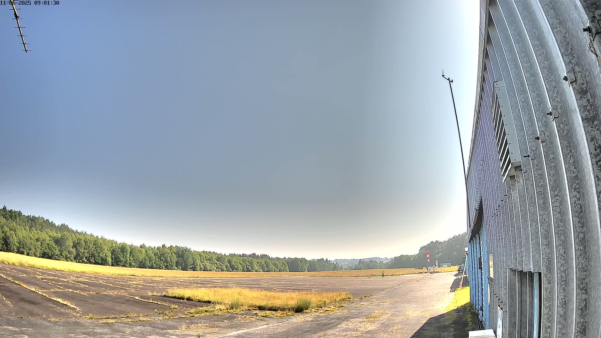 A wide, flat, paved area, possibly an airfield, stretches out before a line of trees under a clear, bright blue sky, with a corrugated metal building visible at the right.