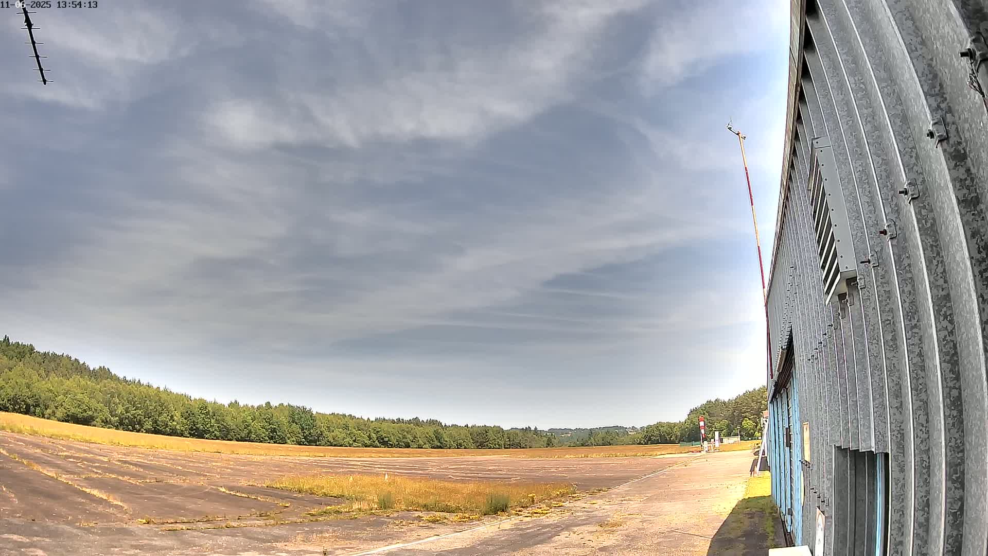 A partly cloudy sky overlooks a flat, overgrown airfield next to a corrugated metal building.