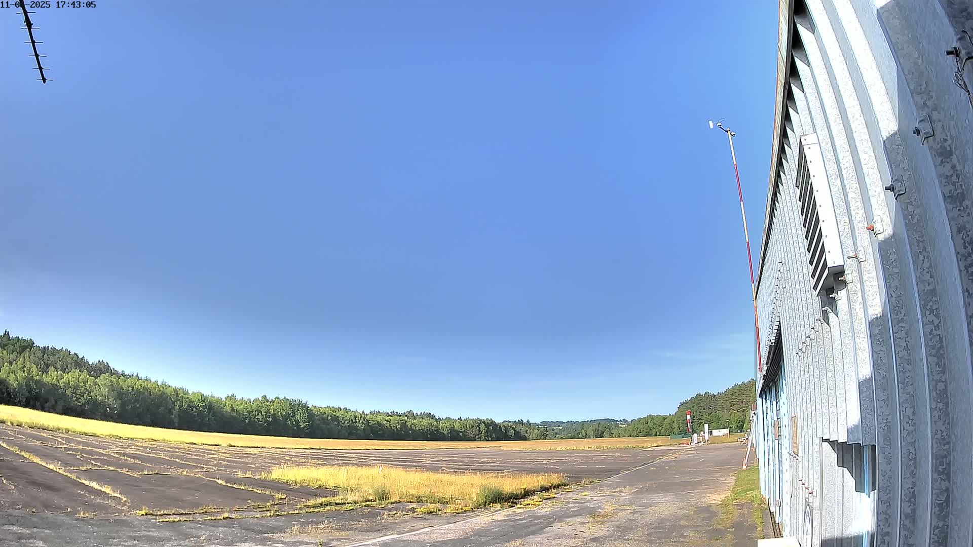 A wide, paved area, possibly an airfield, stretches before a line of trees under a clear blue sky, with a corrugated metal building on the right.