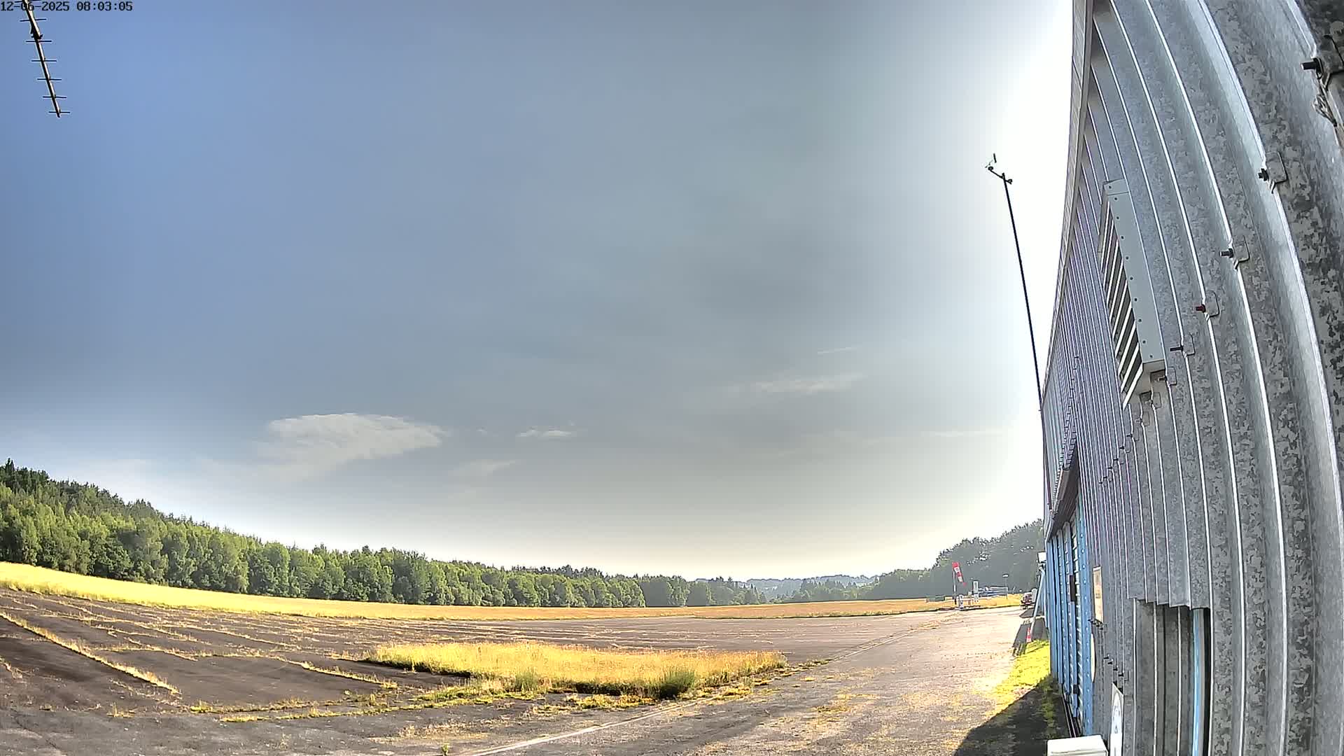 A mostly clear sky above a flat, grassy field with a paved area, next to a corrugated metal building under a mostly sunny sky.