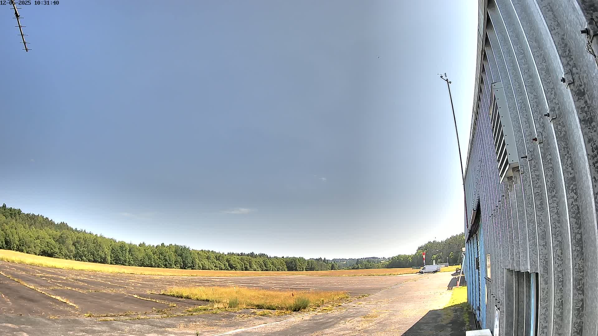 A large, empty paved area, possibly a former airfield, is bordered by trees under a clear, sunny sky, with a corrugated metal building visible to the right.