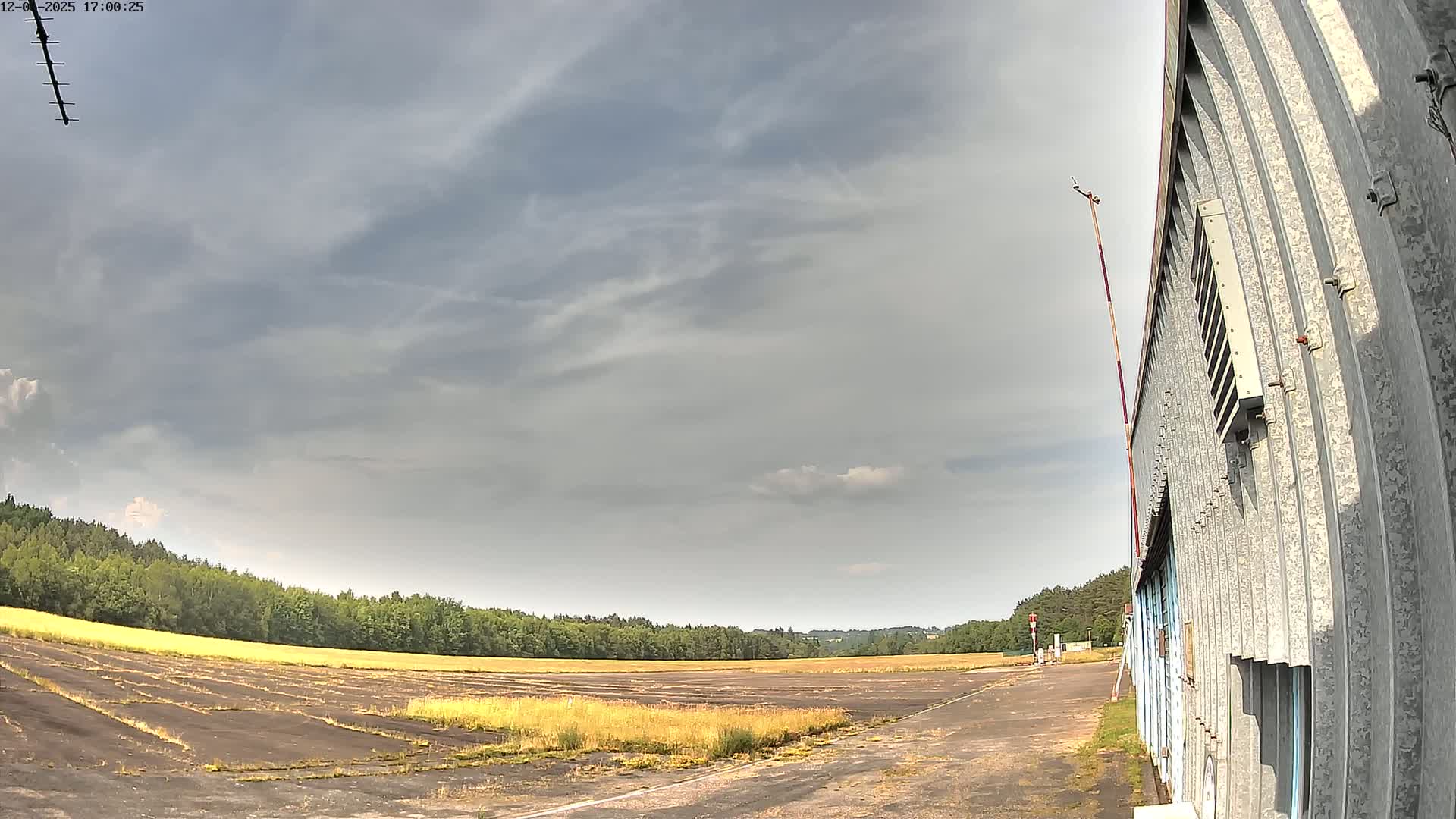A mostly cloudy sky hangs over a flat, grassy field next to a long, corrugated metal building.