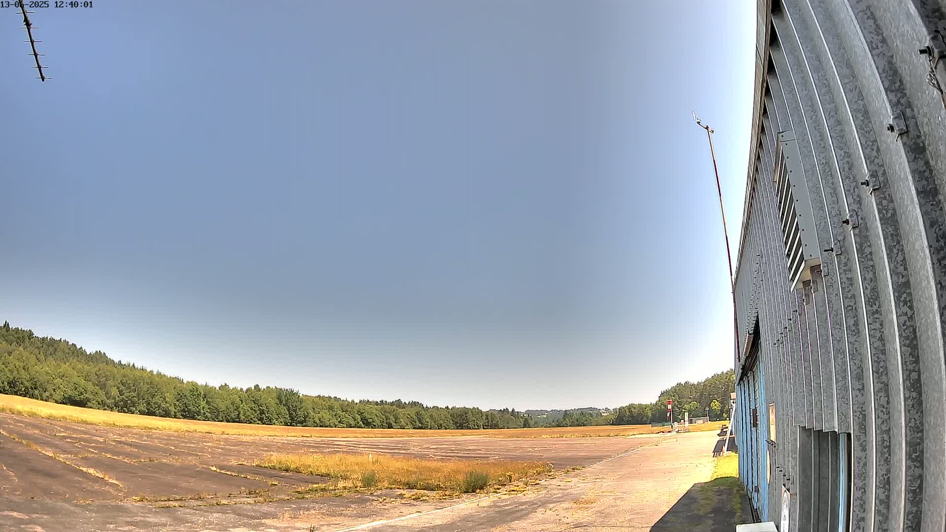 A wide, flat, paved area, possibly a runway or airfield, bordered by a line of trees under a clear blue sky, with a corrugated metal building visible on the right.