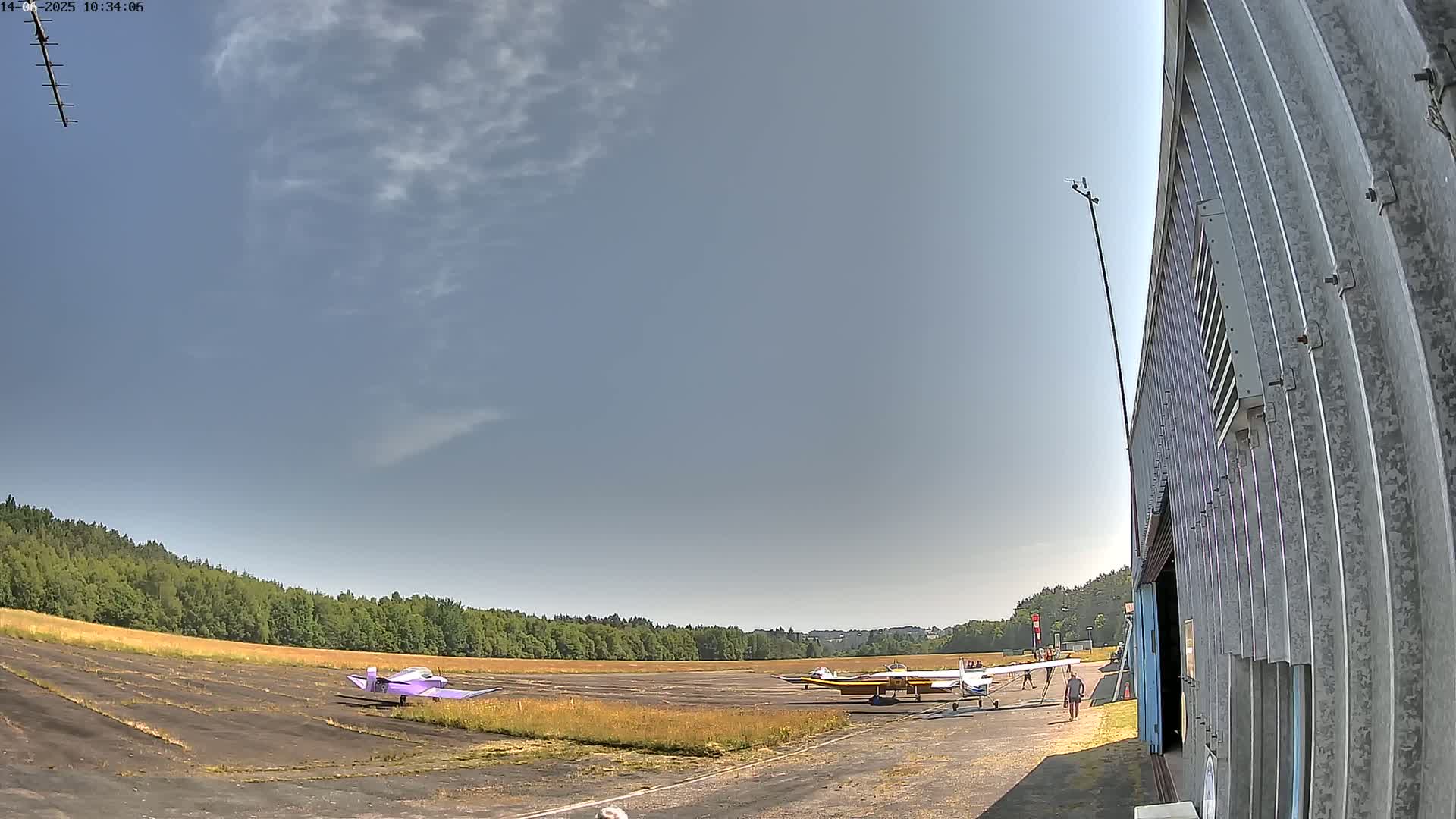 Two small airplanes are parked on a grassy airfield under a mostly clear sky, near a hangar.