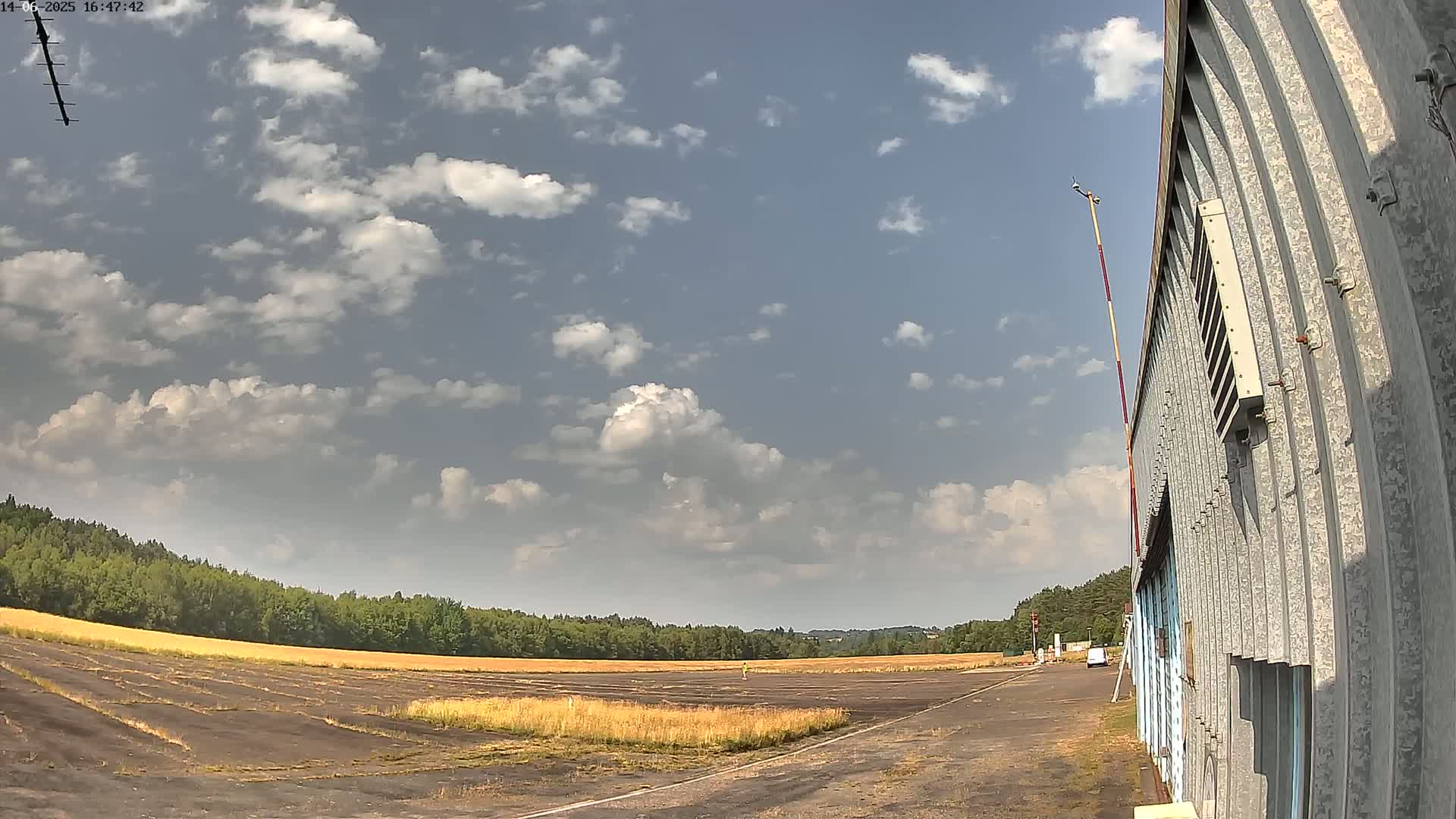 A partly cloudy sky overlooks a flat, grassy airfield next to a corrugated metal building.