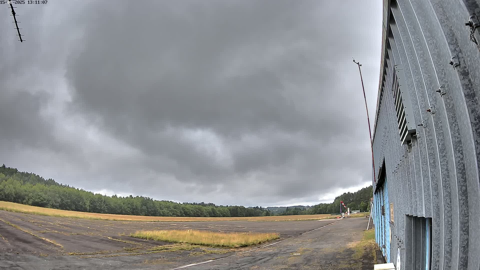 A gray, overcast sky looms over a flat, paved area next to a corrugated metal building, with a line of trees in the distance.