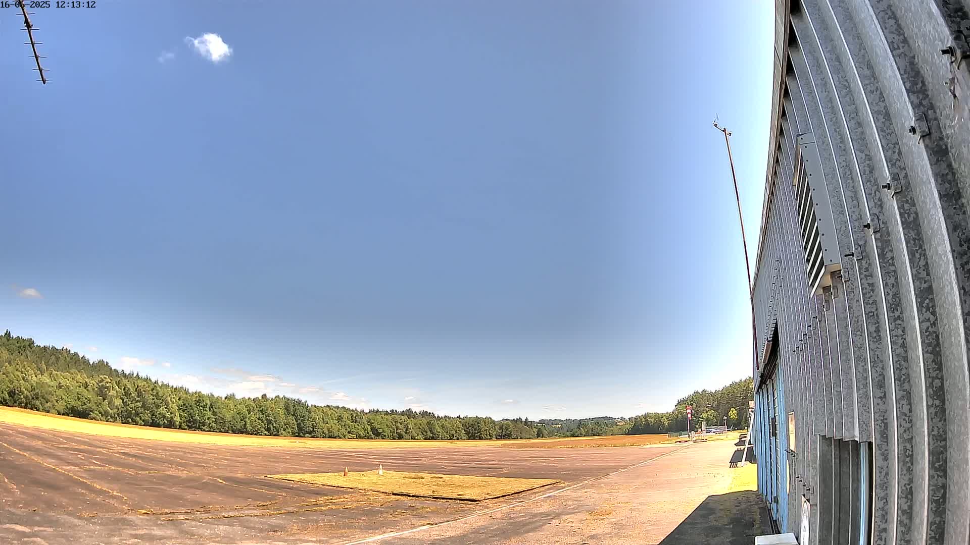 A mostly clear blue sky is over a flat, paved area bordered by trees, next to a corrugated metal building.