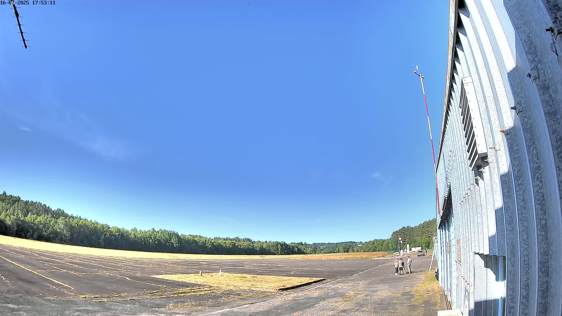A wide, flat, paved area, possibly an airfield, stretches out under a clear blue sky, with a few people standing near a corrugated metal building in the foreground, and a line of trees in the background.