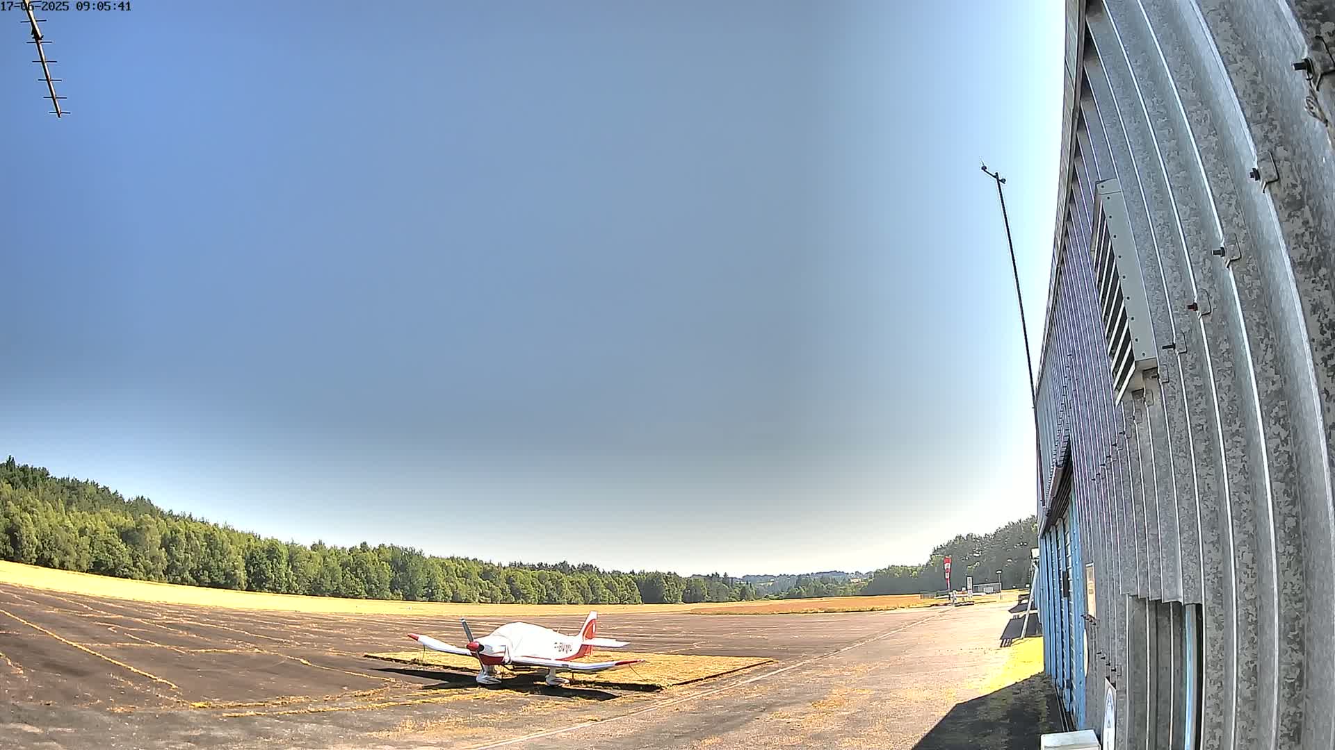 A small, single-engine airplane sits on a tarmac next to a corrugated metal building under a clear, sunny sky.