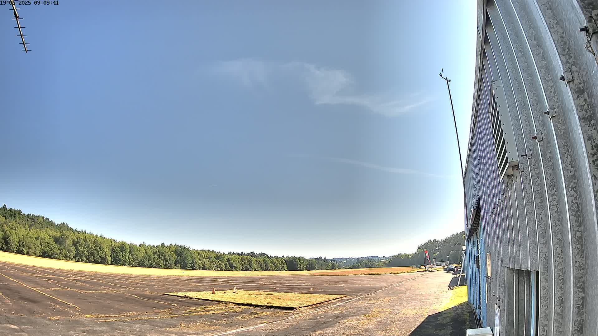 A wide, mostly empty paved area, bordered by trees and a corrugated metal building, under a mostly clear blue sky with a few wispy clouds.
