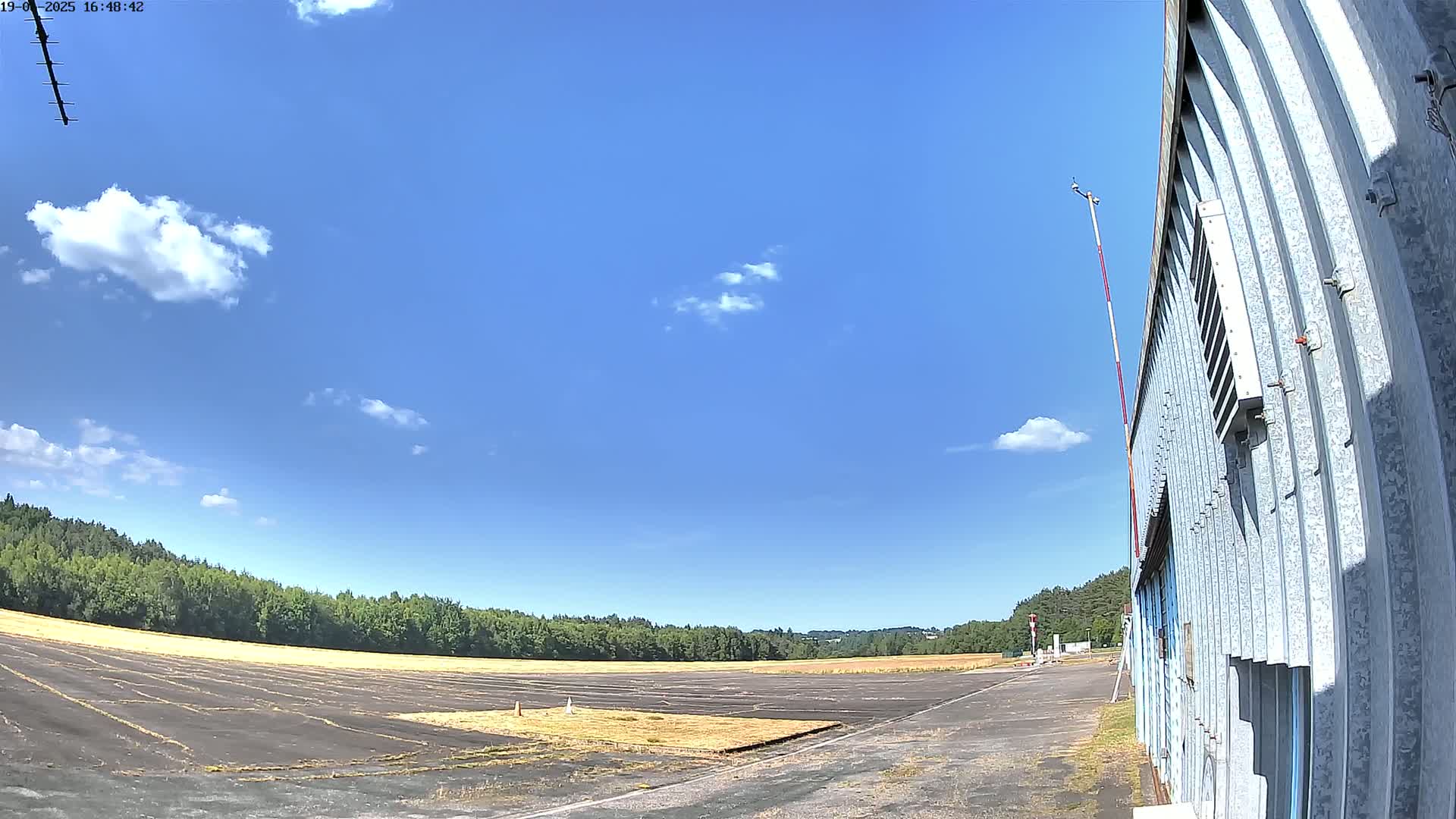 A mostly sunny day shows a paved airstrip next to a corrugated metal building, with a line of trees in the background.