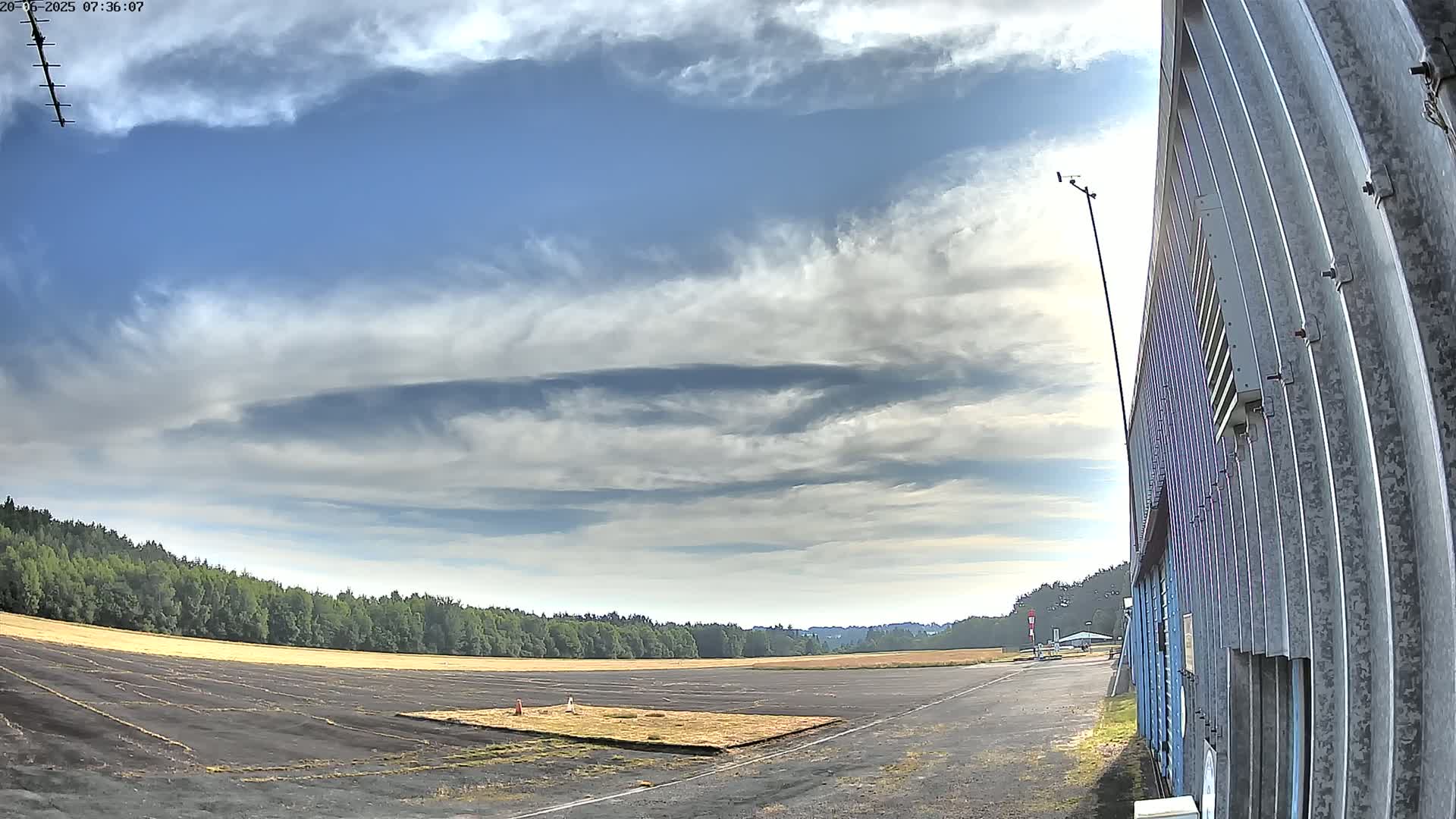 A paved runway extends into a field bordered by trees under a partly cloudy sky.
