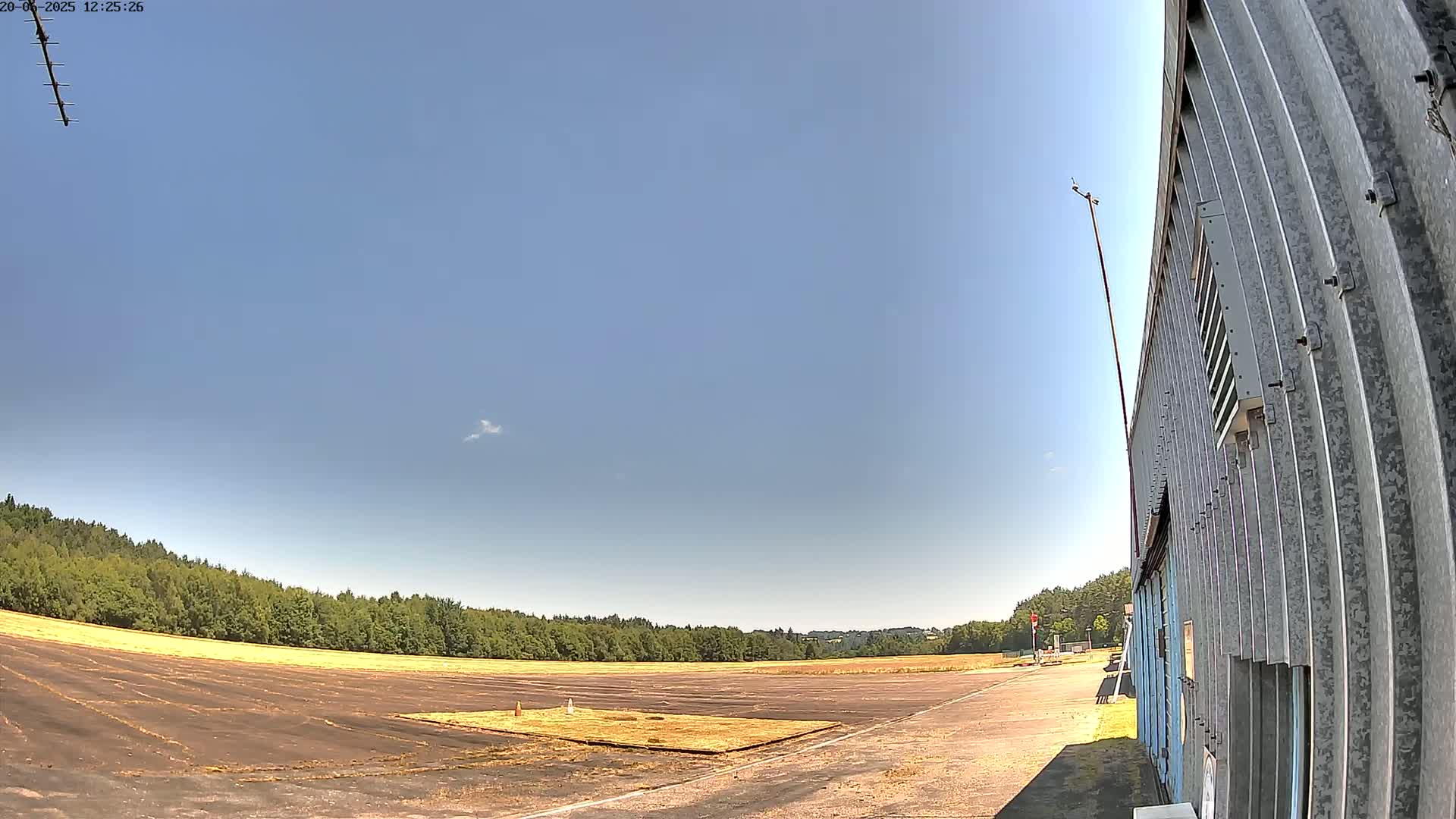 A large, empty paved area bordered by trees under a mostly clear, sunny sky next to a corrugated metal building.