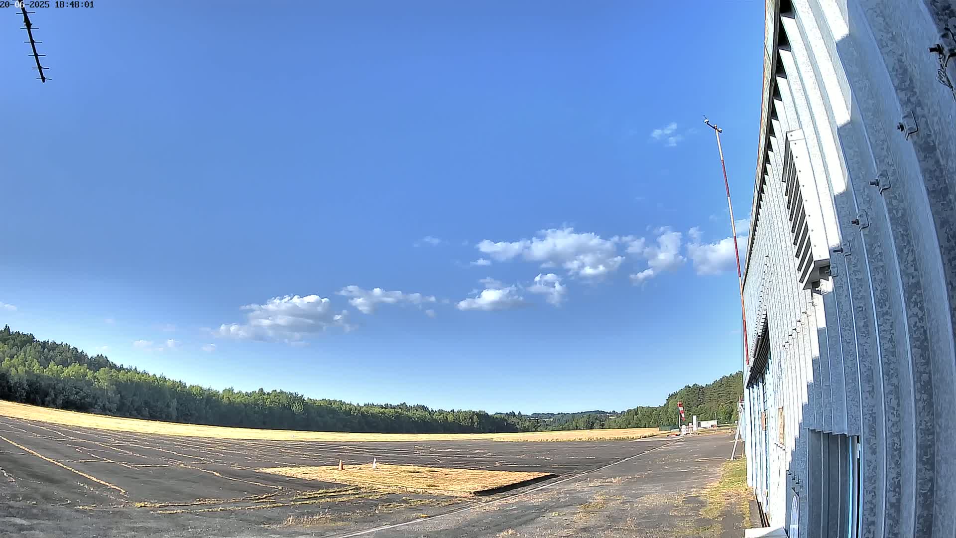 A mostly clear blue sky with scattered cumulus clouds overlooks a flat, paved airstrip bordered by trees, adjacent to a corrugated metal building.