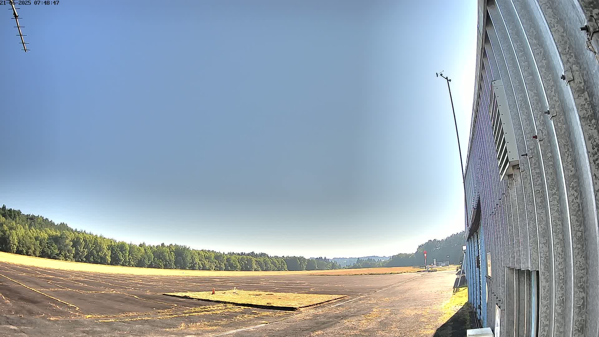 A mostly clear sky overlooks a flat, paved area bordered by trees, next to a corrugated metal building.