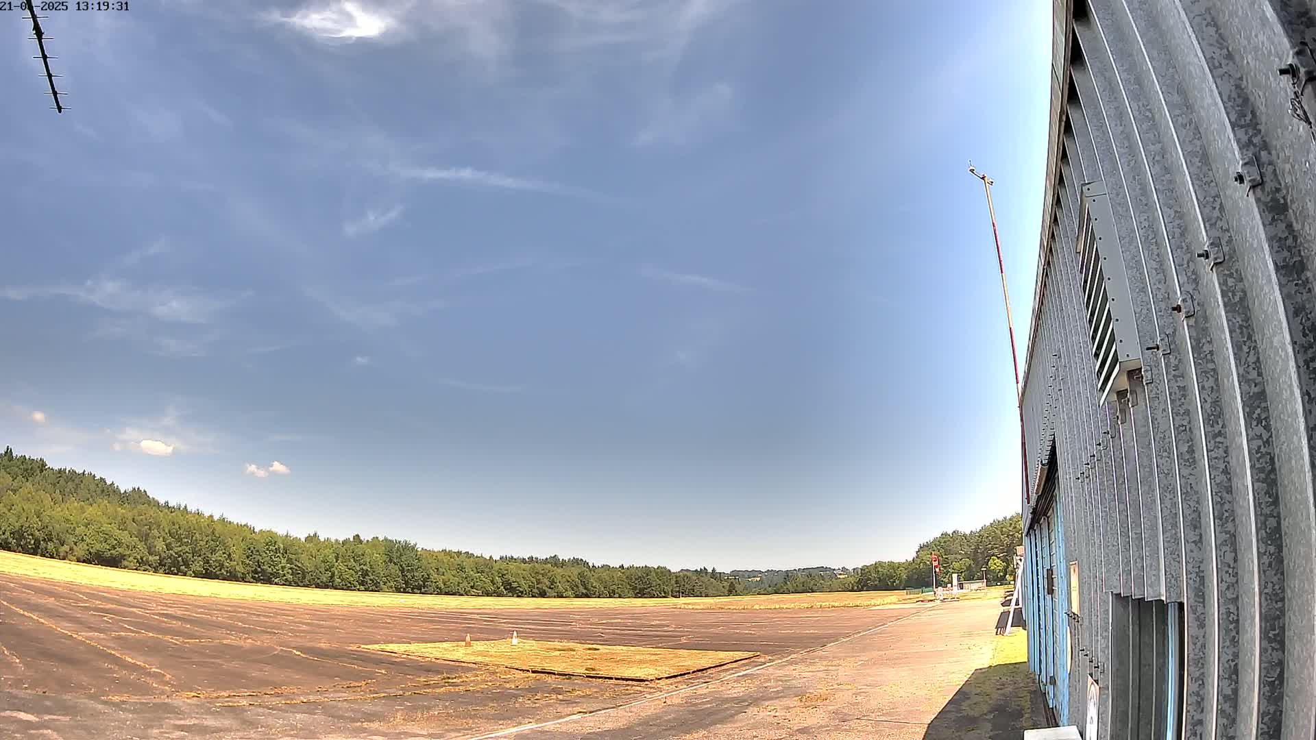 A mostly clear blue sky is over a flat, brown airfield bordered by a line of green trees and a corrugated metal building.